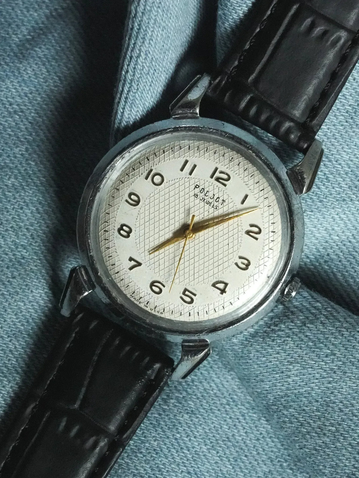 Vintage Poljot watch with white textured dial, black leather strap, gold hands on denim