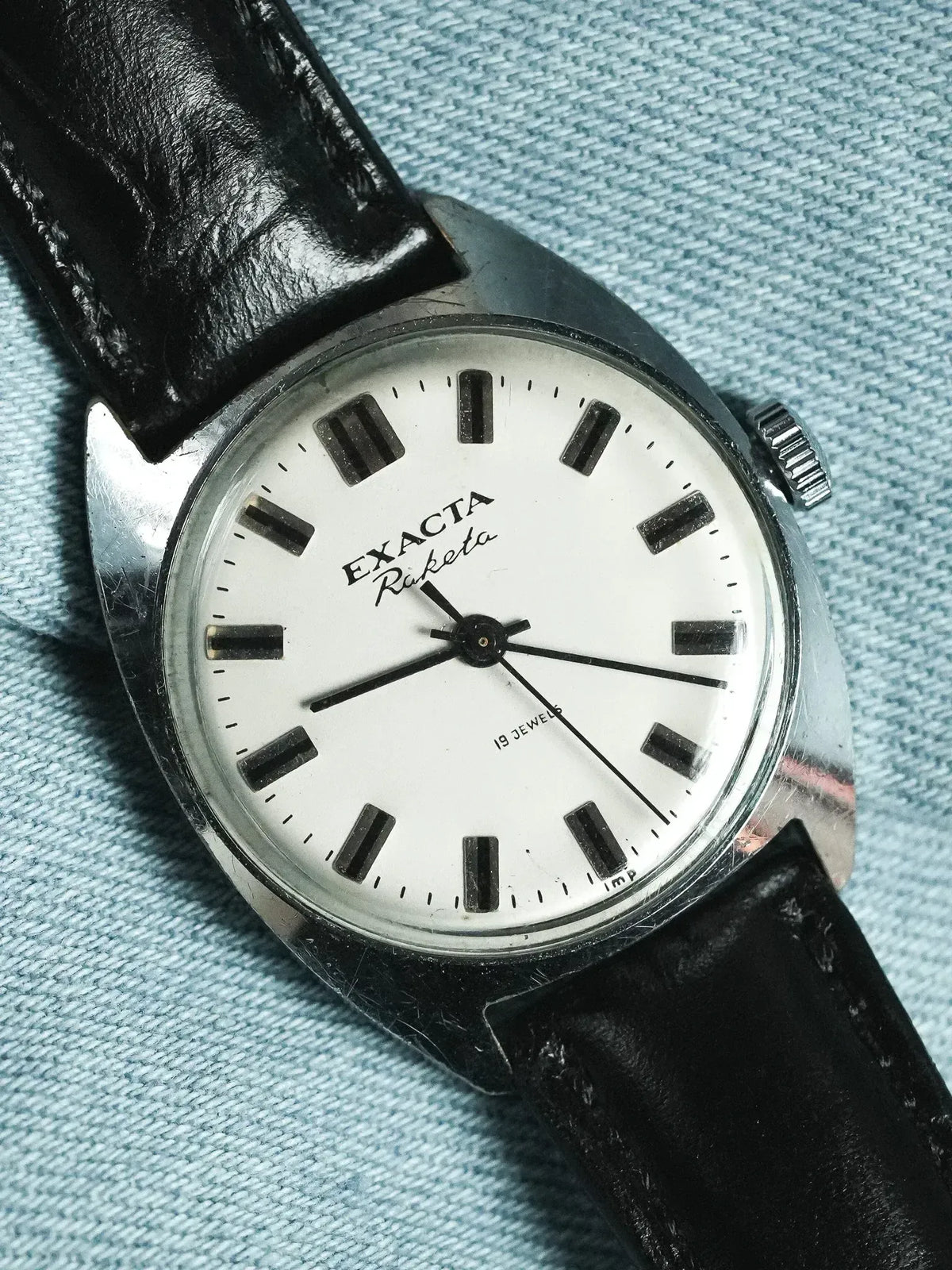 Exacta Raketa vintage watch with white dial, black markers, and black leather strap on denim