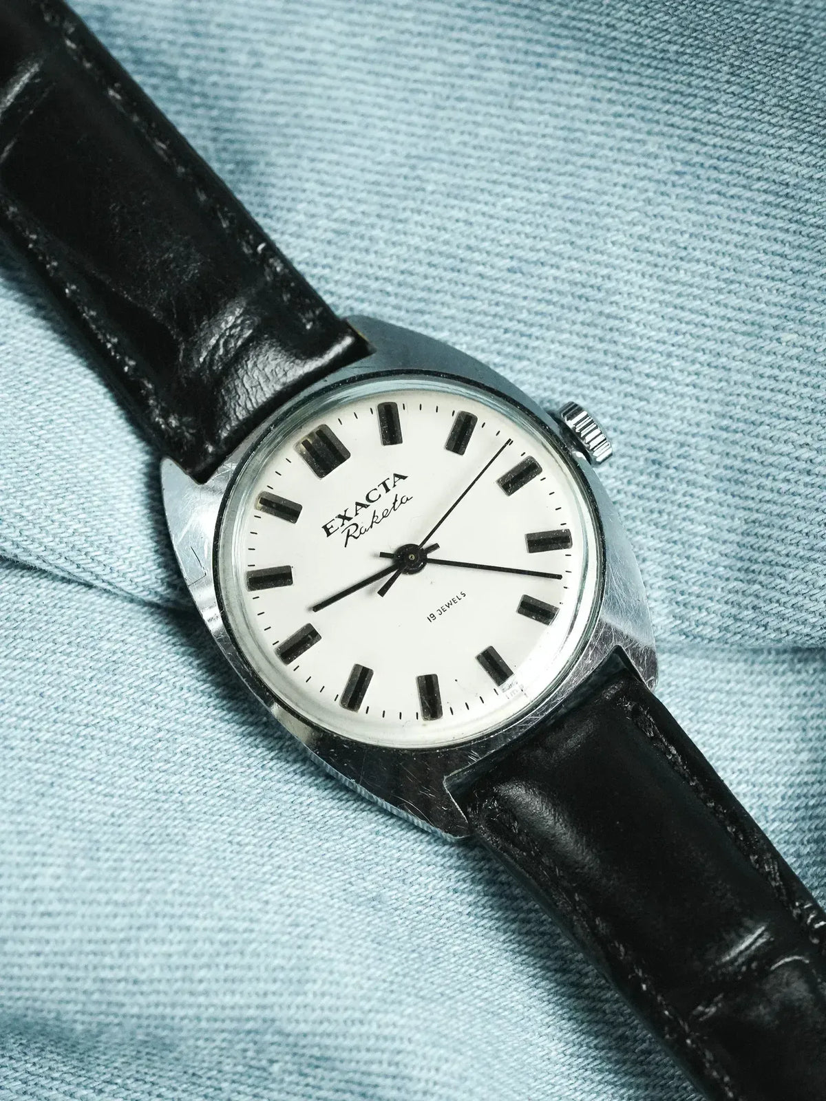 Exacta Riketa vintage watch with white dial, black markers, and black leather strap on denim