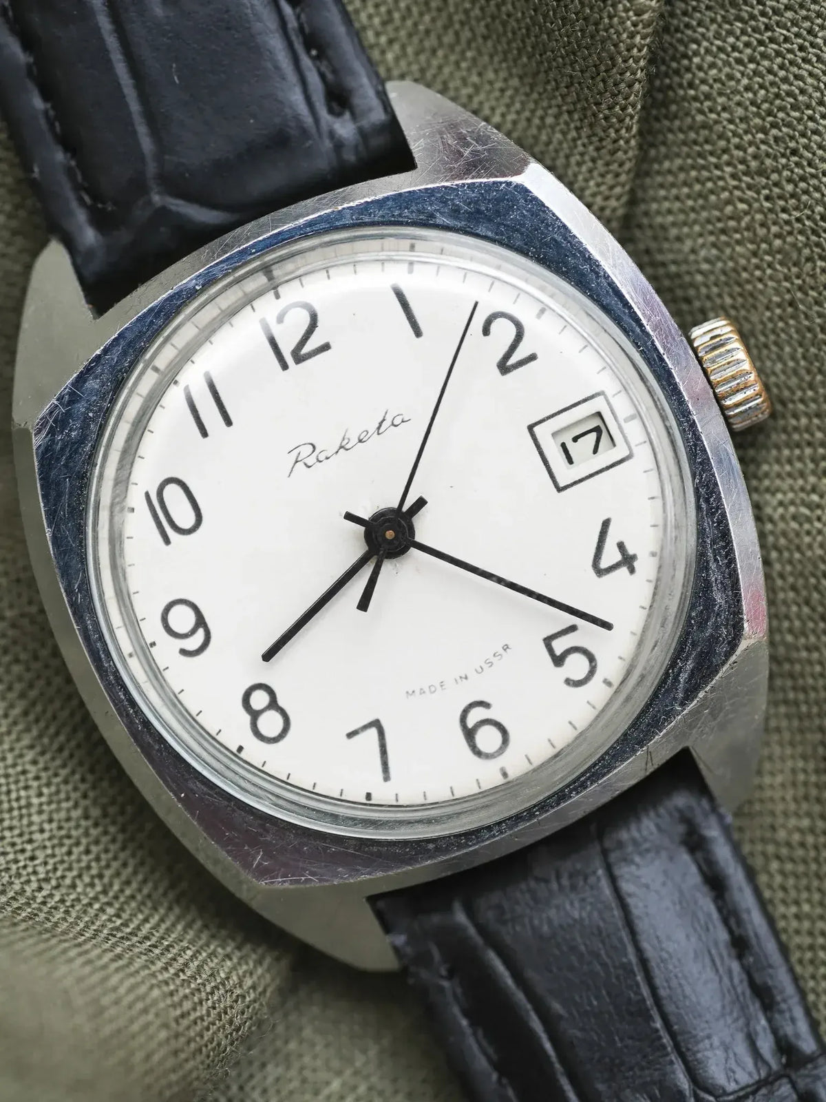Raketa vintage watch with white dial, black leather strap, and date window on olive fabric