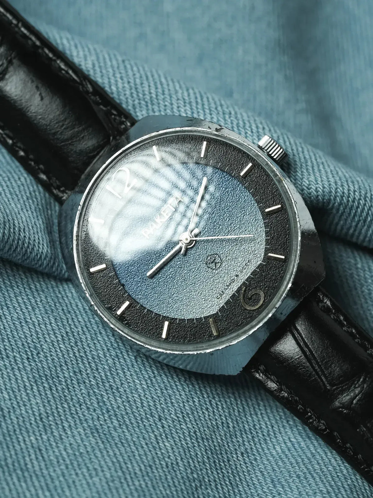 Vintage Raketa watch with blue textured dial, black leather strap, on denim background