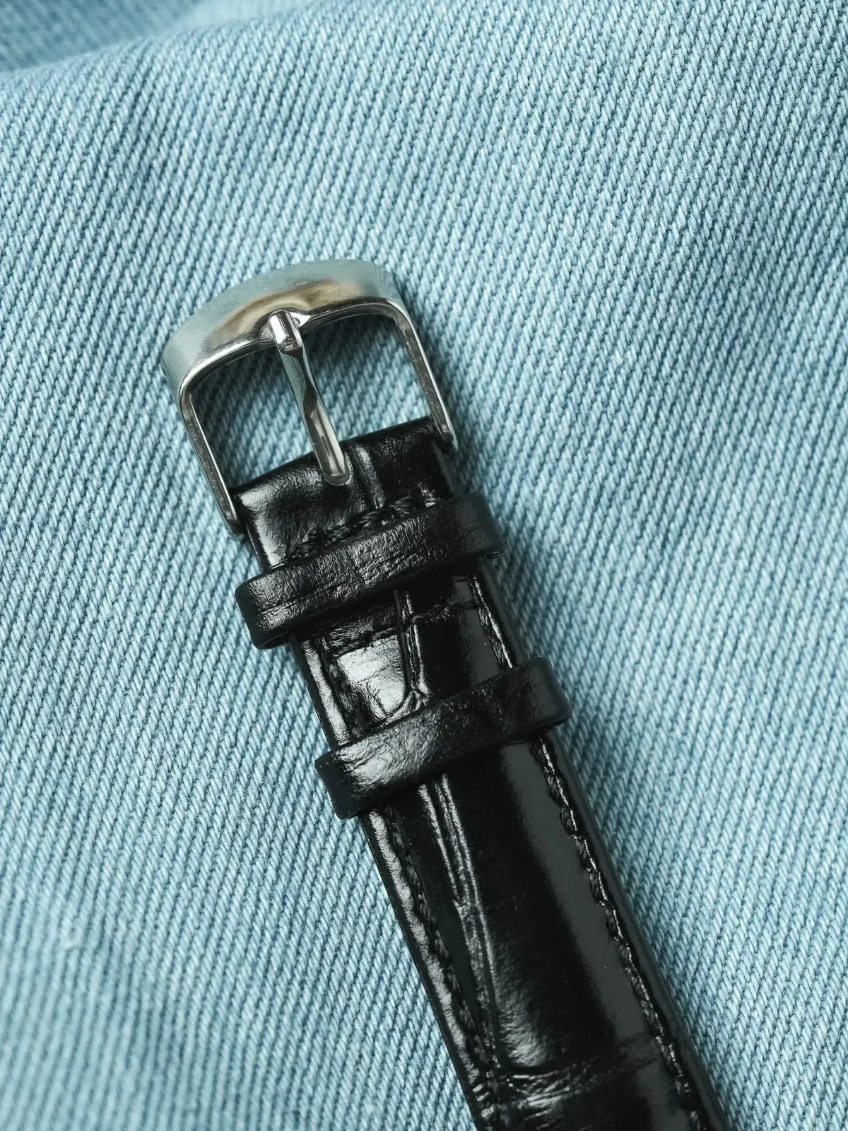Black alligator leather watch strap with silver buckle on denim background