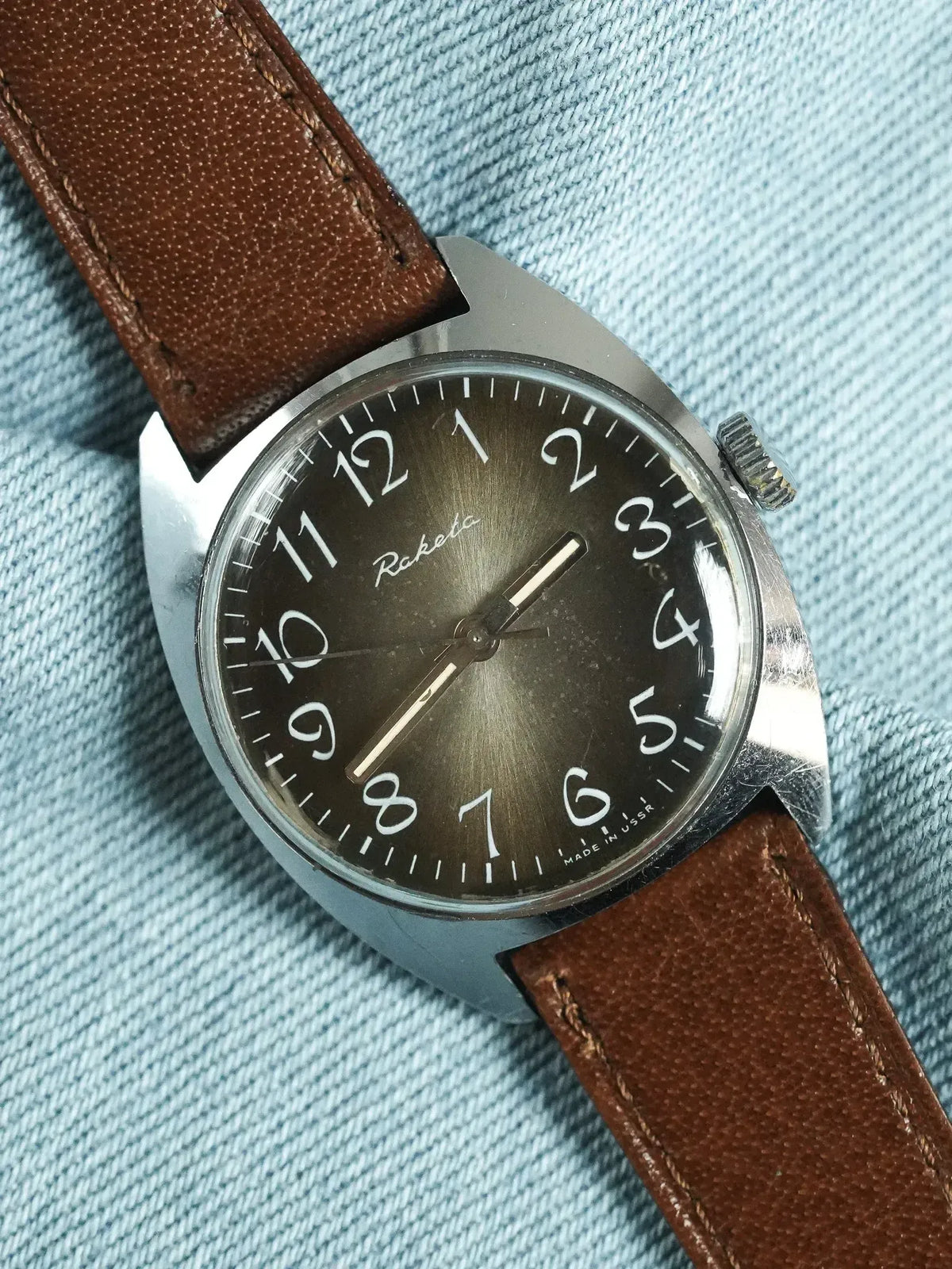 Raketa vintage watch with brown leather strap, black dial, and steel case on denim