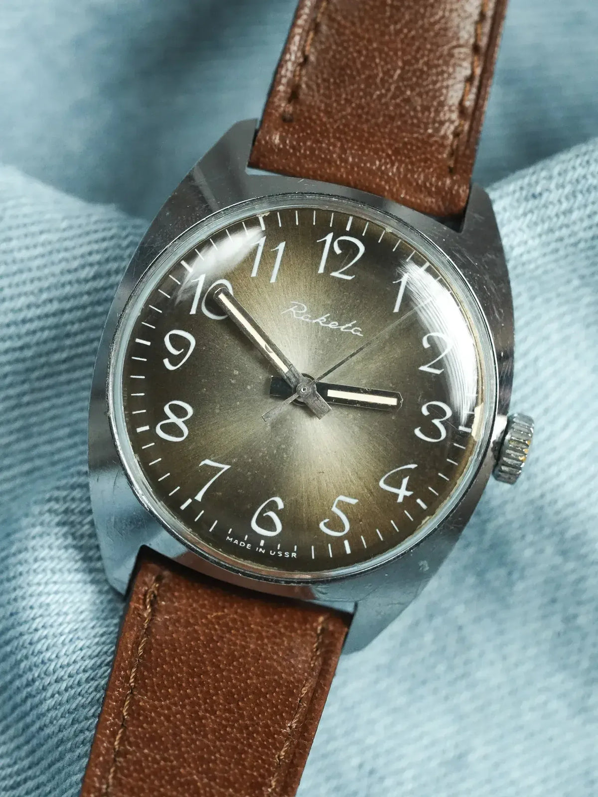 Raketa vintage watch with brown leather strap, silver case, and dark dial on denim