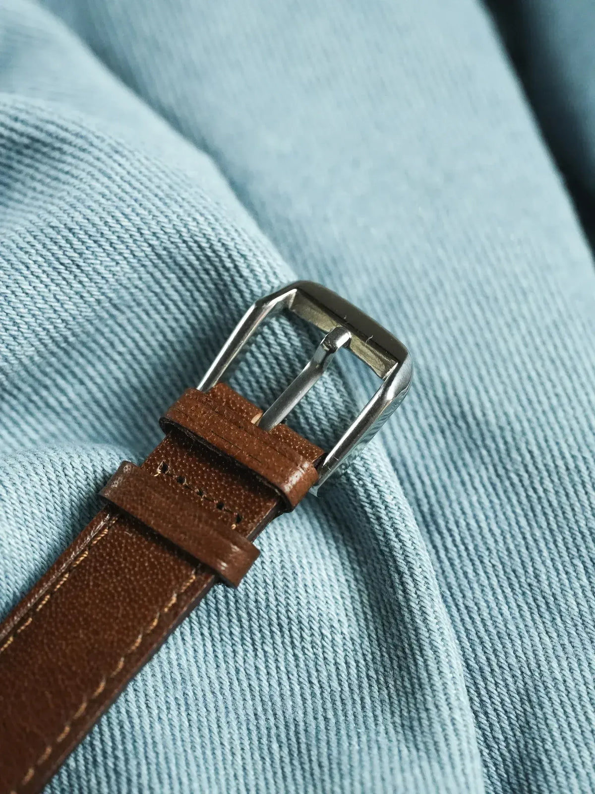 Brown leather watch strap with silver buckle on light blue fabric
