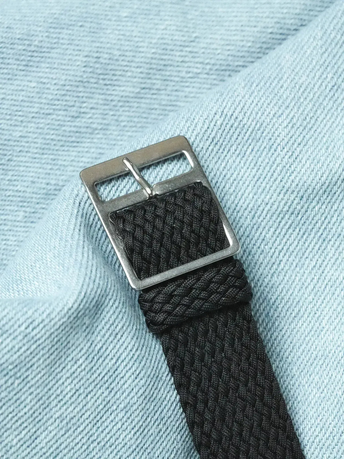 Black braided nylon watch strap with silver-toned buckle on light blue fabric