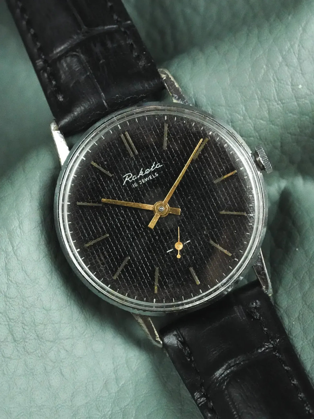 Raketa vintage watch with black textured dial, gold hands, and black leather strap
