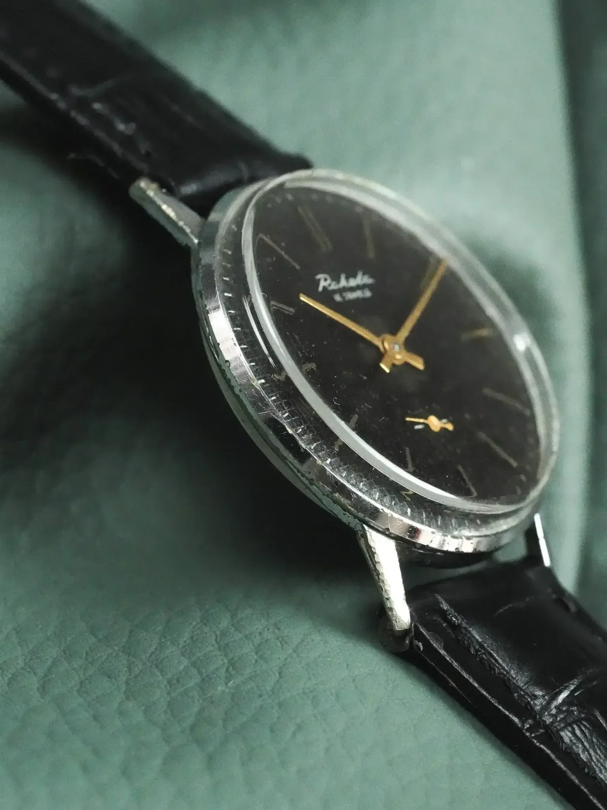 Vintage Raketa watch with black dial, gold hands, and black leather strap on green background
