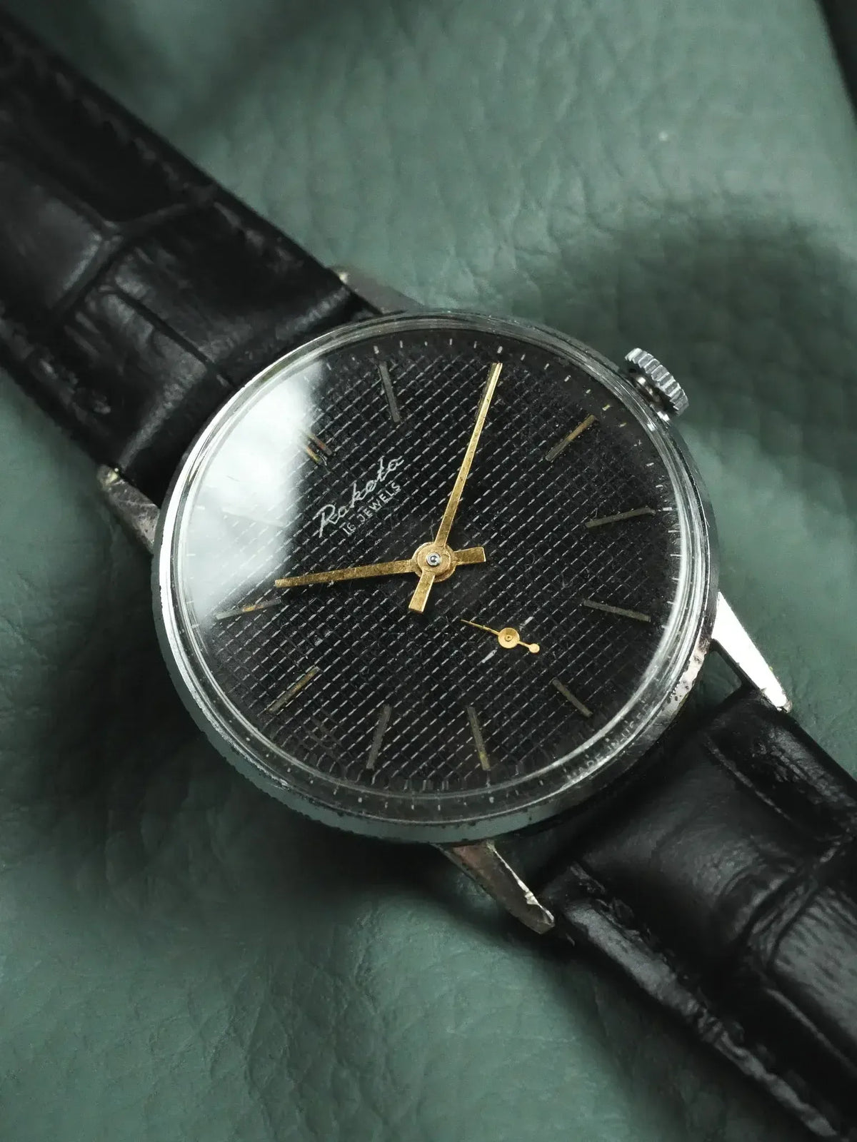 Raketa vintage watch with black textured dial, gold hands, and black leather strap on green background