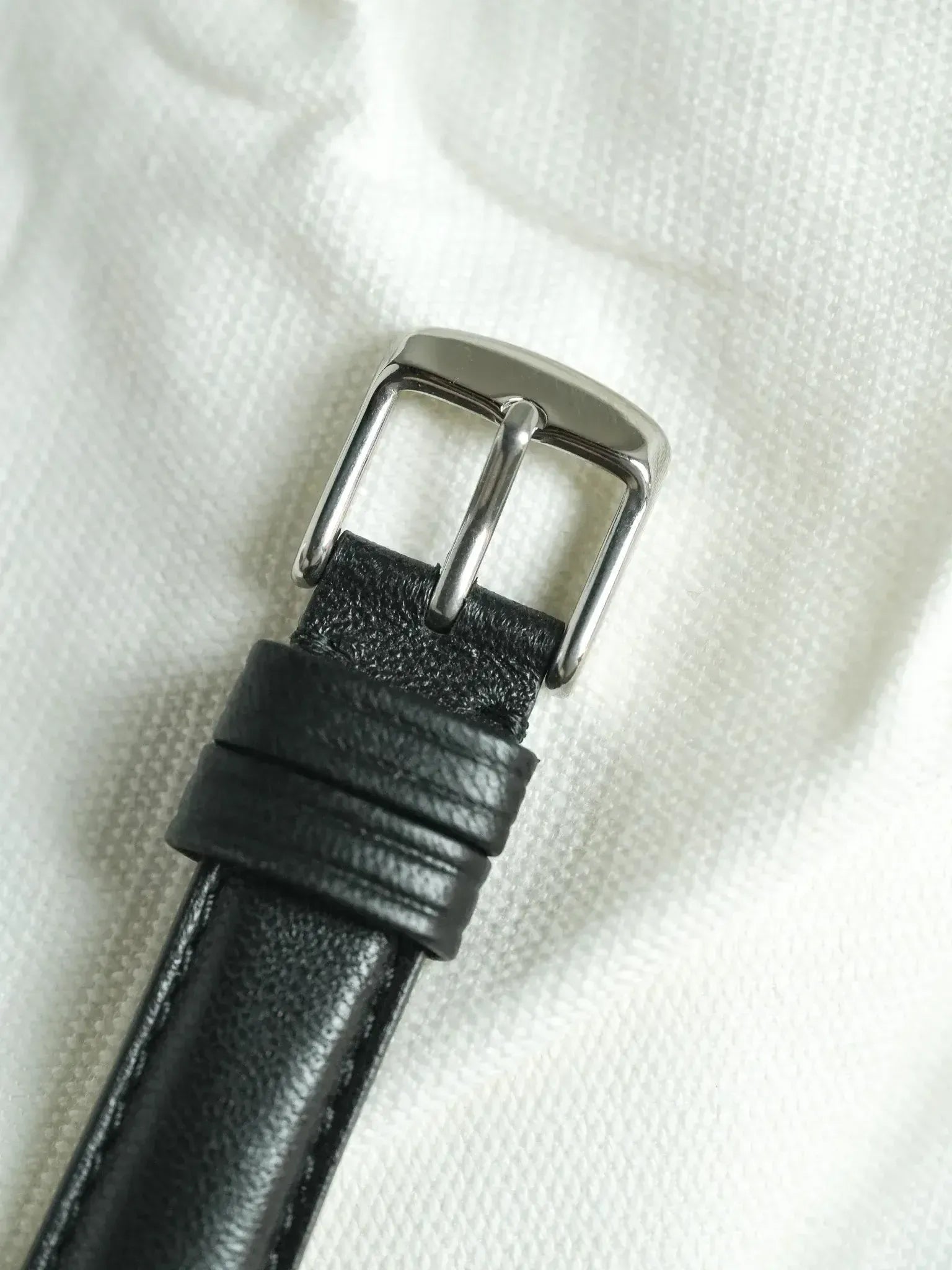 Black leather watch strap with polished steel buckle on white fabric