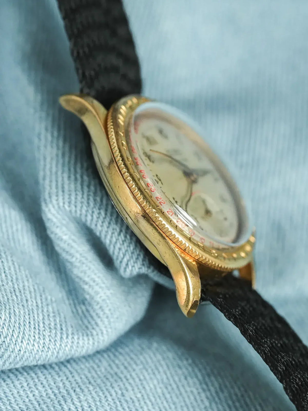 Vintage gold watch with textured bezel, cream dial, red numerals, black strap on blue fabric