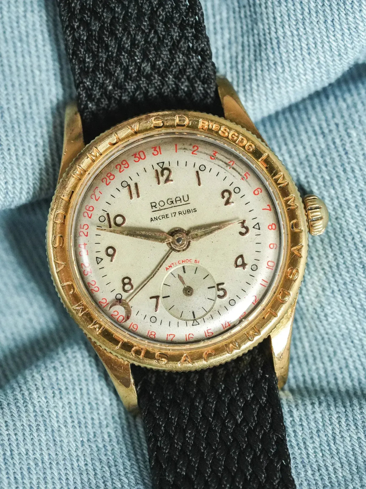 Vintage Rogau watch with gold case, cream dial, red date, black fabric strap on denim