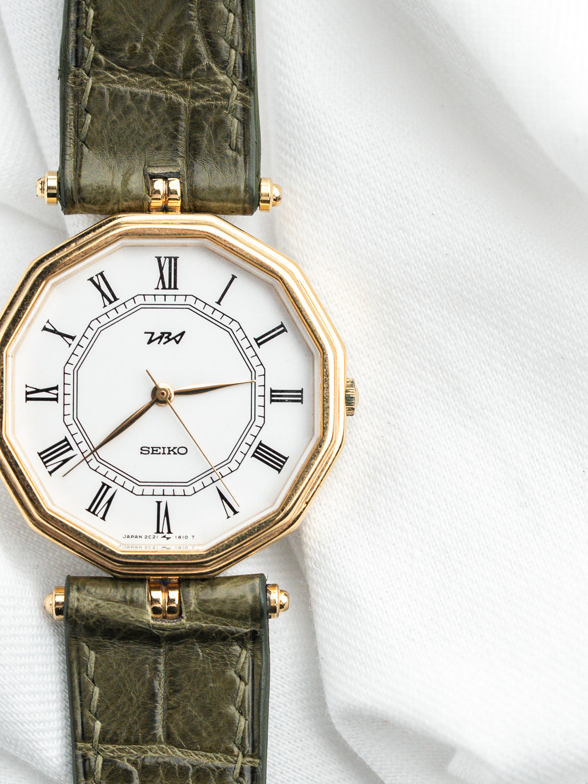 Montre Seiko vintage avec cadran blanc, boîtier doré octogonal et bracelet en cuir vert.