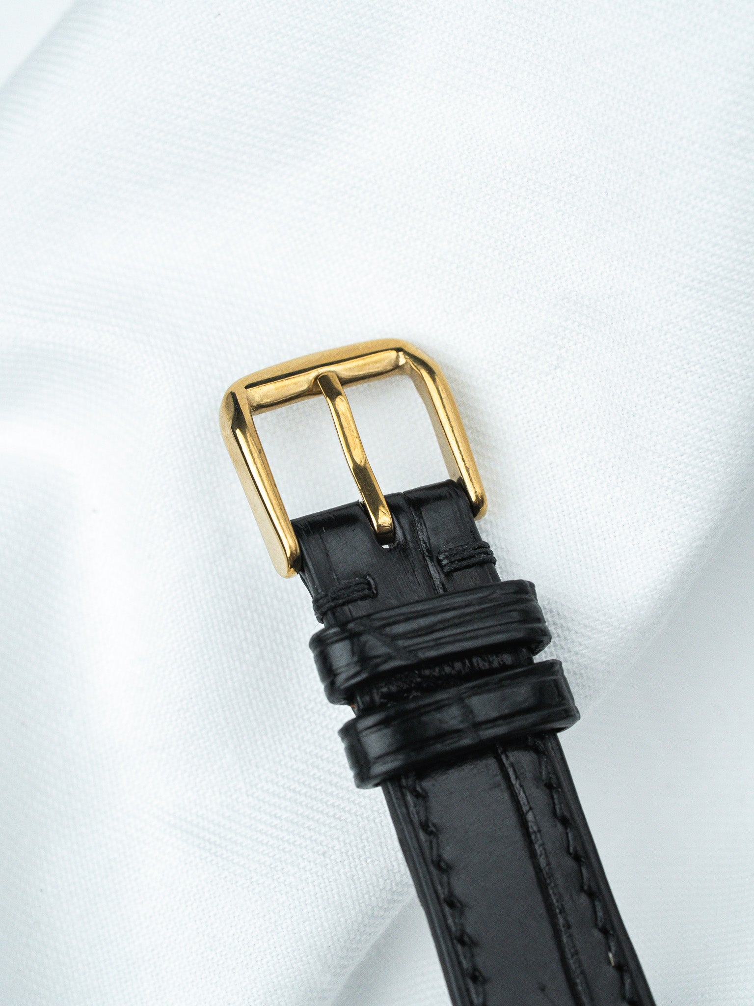 Black leather watch strap with gold buckle on white fabric background