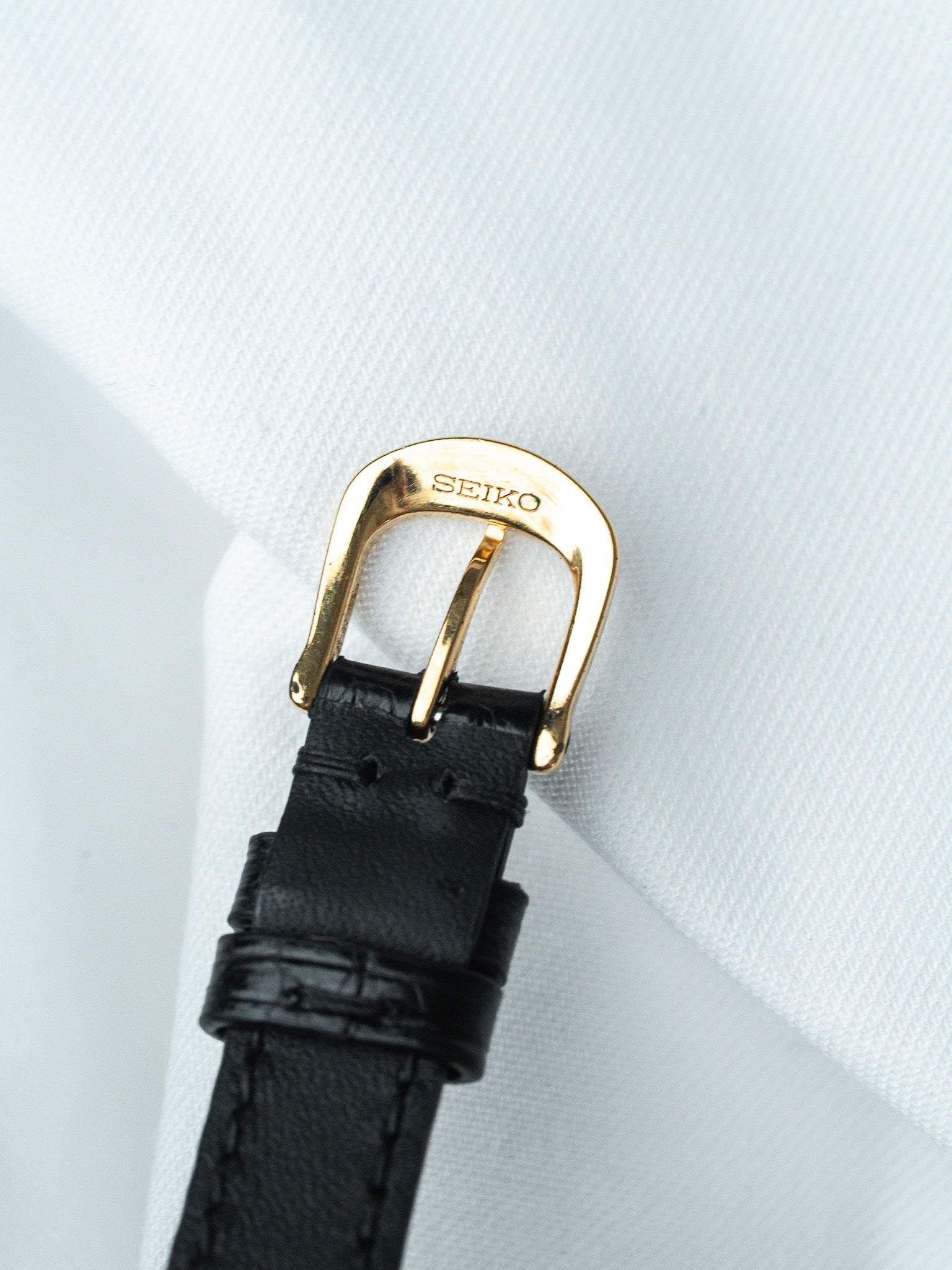 Seiko watch buckle with gold-toned clasp and black leather strap on white fabric