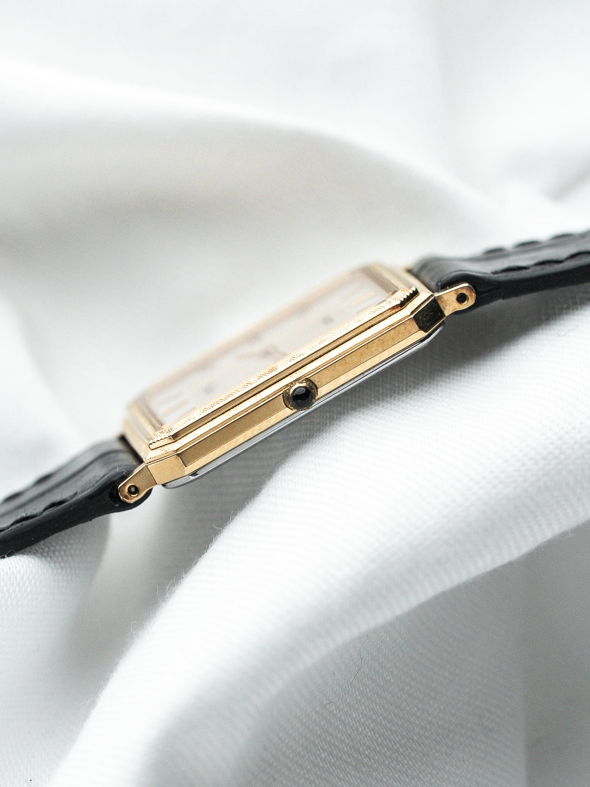 Montre vintage au boîtier rectangulaire doré et bracelet en cuir noir sur fond blanc