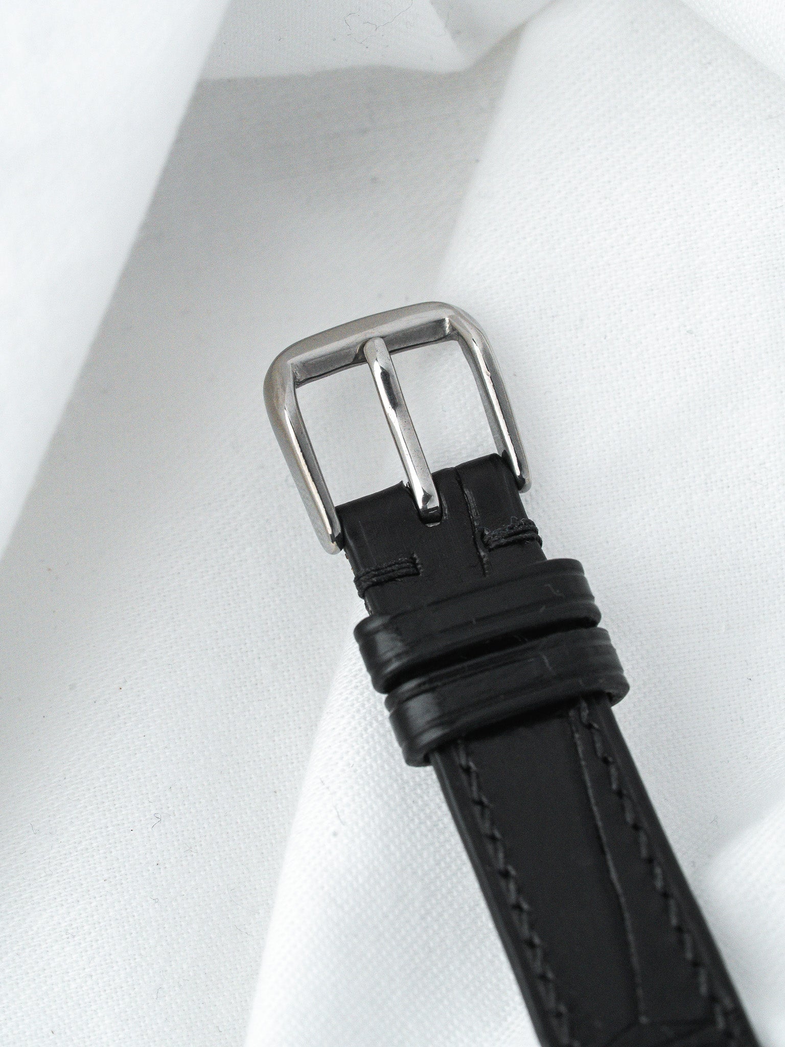 Black leather watch strap with silver buckle, stitched edges, on white fabric