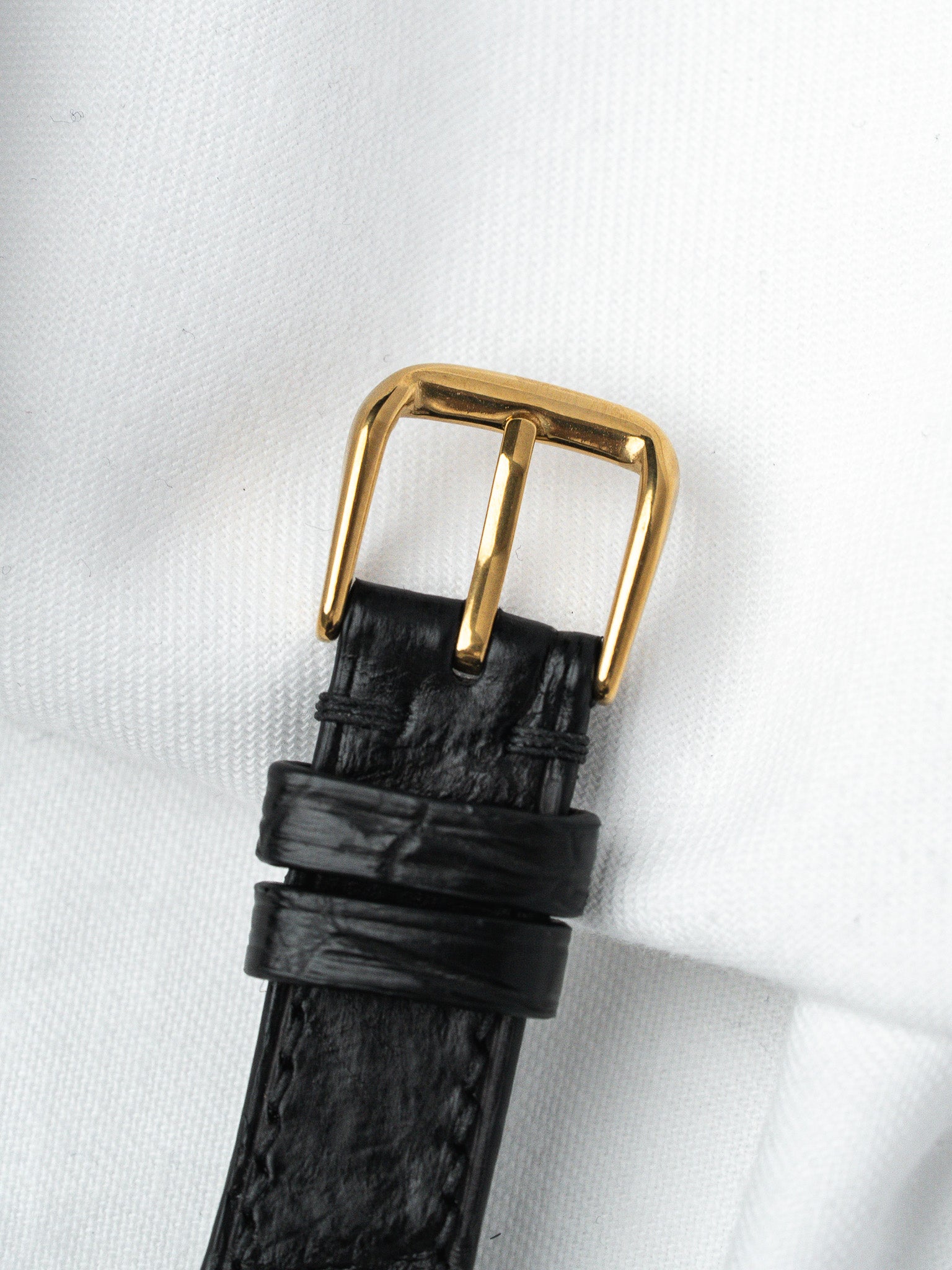Black textured leather watch strap with gold buckle on white fabric