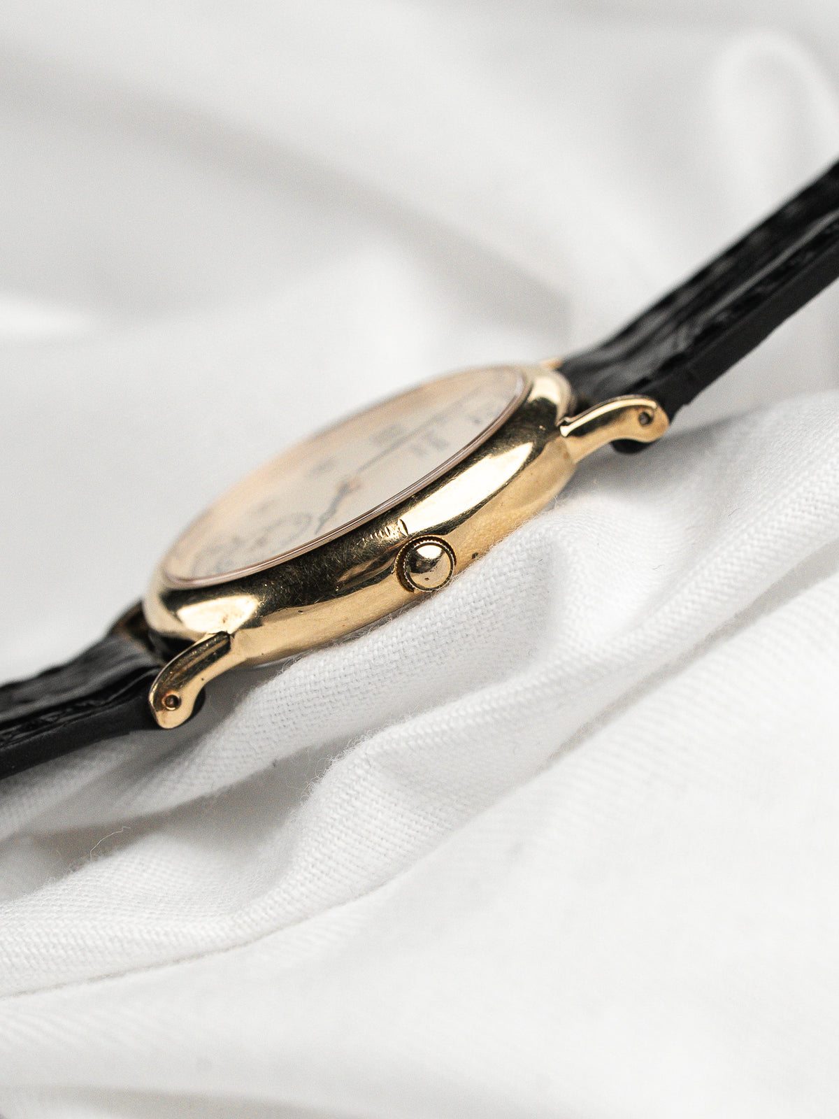 Montre vintage avec boîtier doré et bracelet en cuir noir sur tissu blanc.