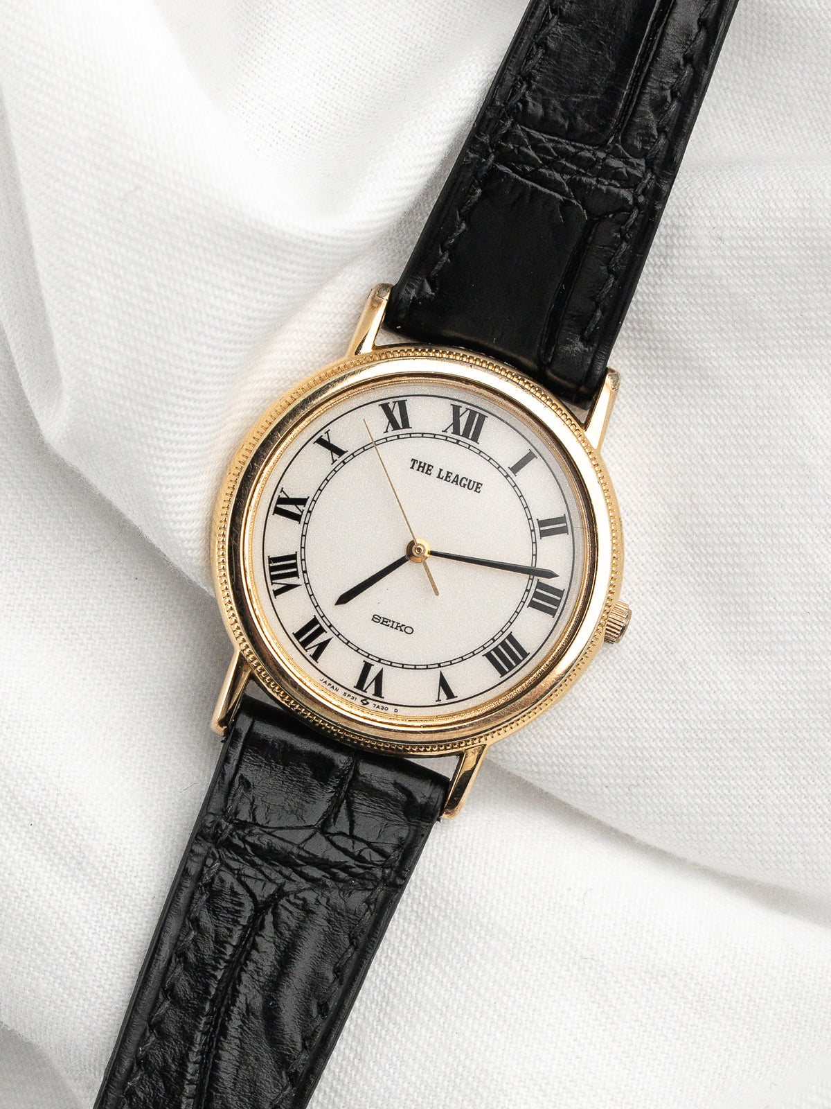 Montre Seiko vintage au boîtier doré, cadran blanc à chiffres romains et bracelet cuir noir.