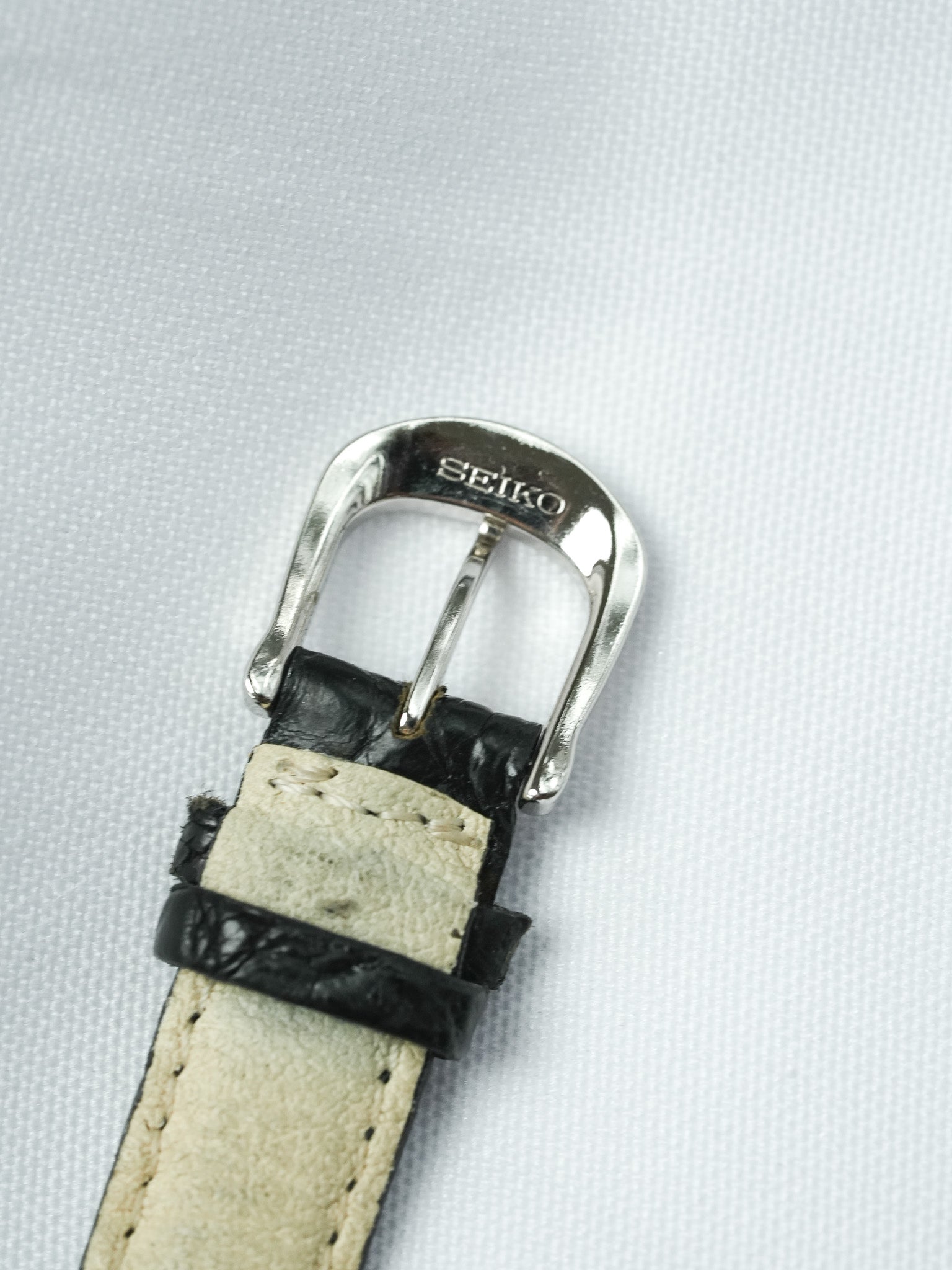 Seiko watch buckle with black leather strap, silver-toned clasp on textured white background