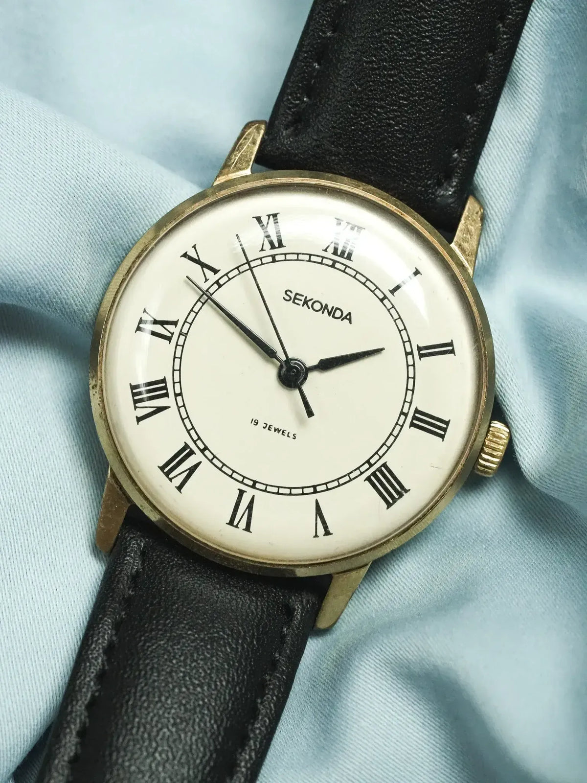 Vintage Sekonda watch with gold case, white dial, Roman numerals, black leather strap on light blue fabric