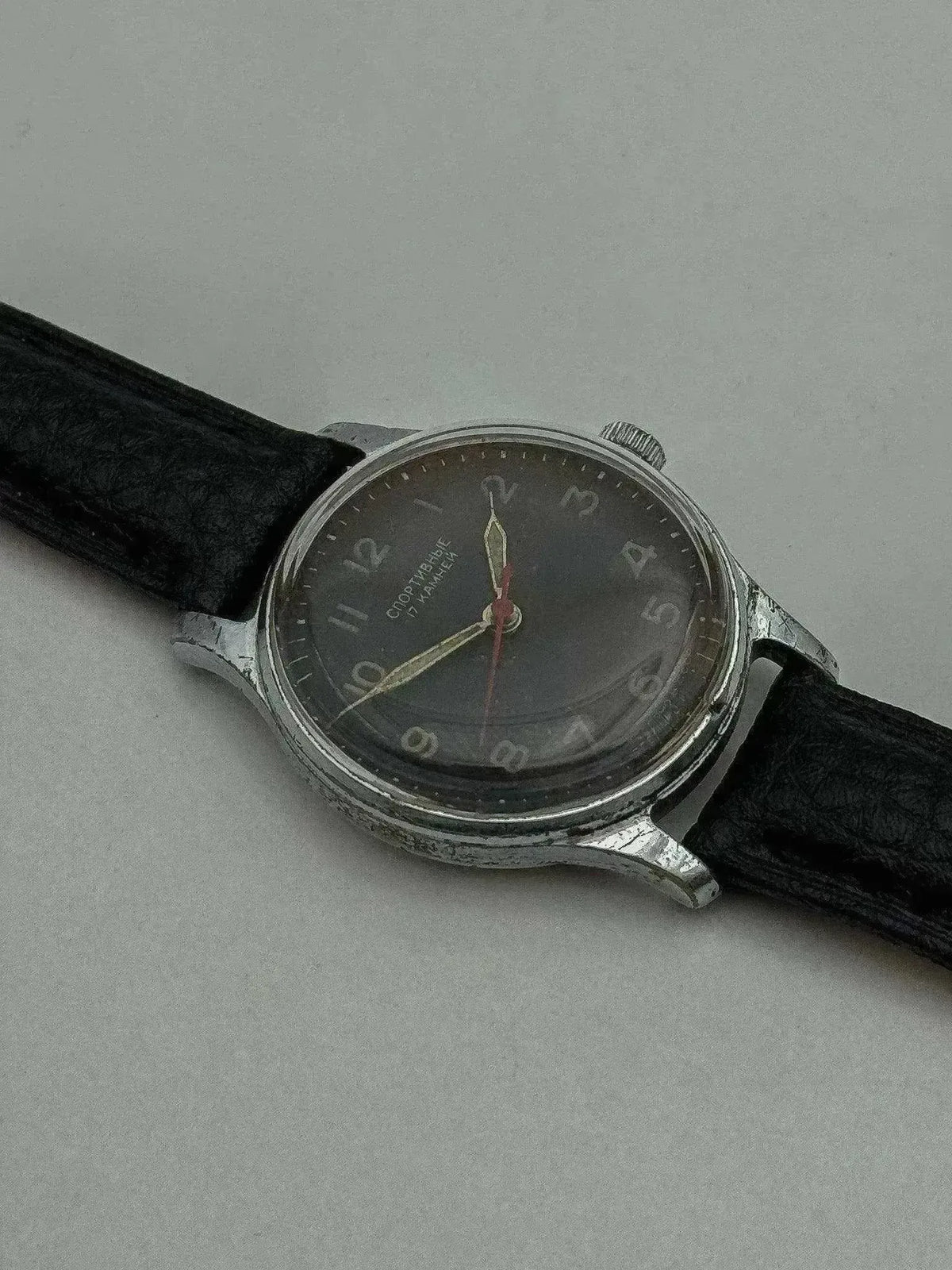 Vintage Soviet Sportivnye watch with dark dial, silver case, black leather strap on neutral background