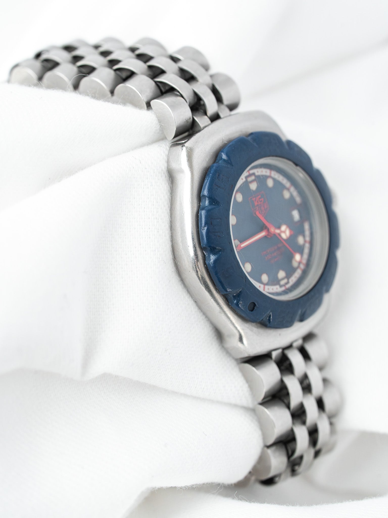 TAG Heuer vintage watch with blue bezel, red hands, and steel bracelet on white fabric