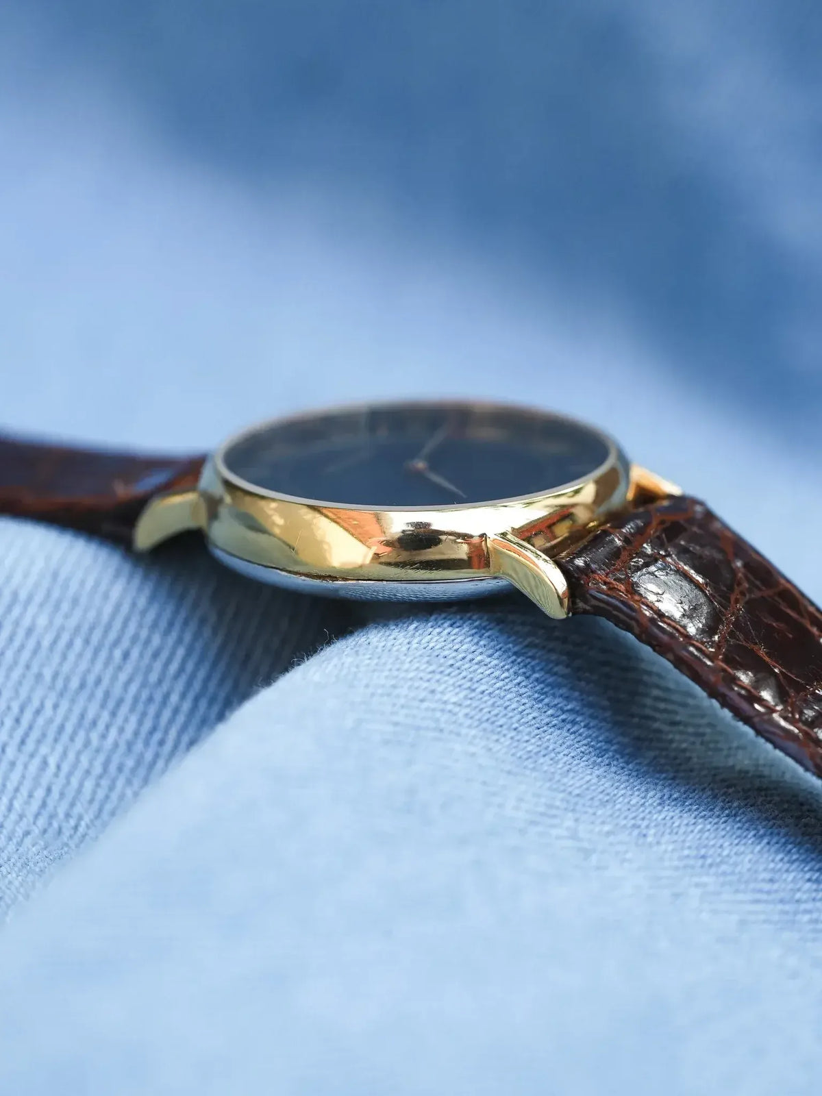 Vintage watch with gold case, black dial, brown alligator strap on blue fabric