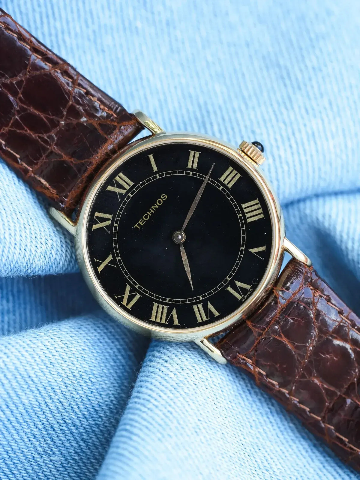 Technos vintage watch with black dial, gold Roman numerals, brown leather strap on blue fabric