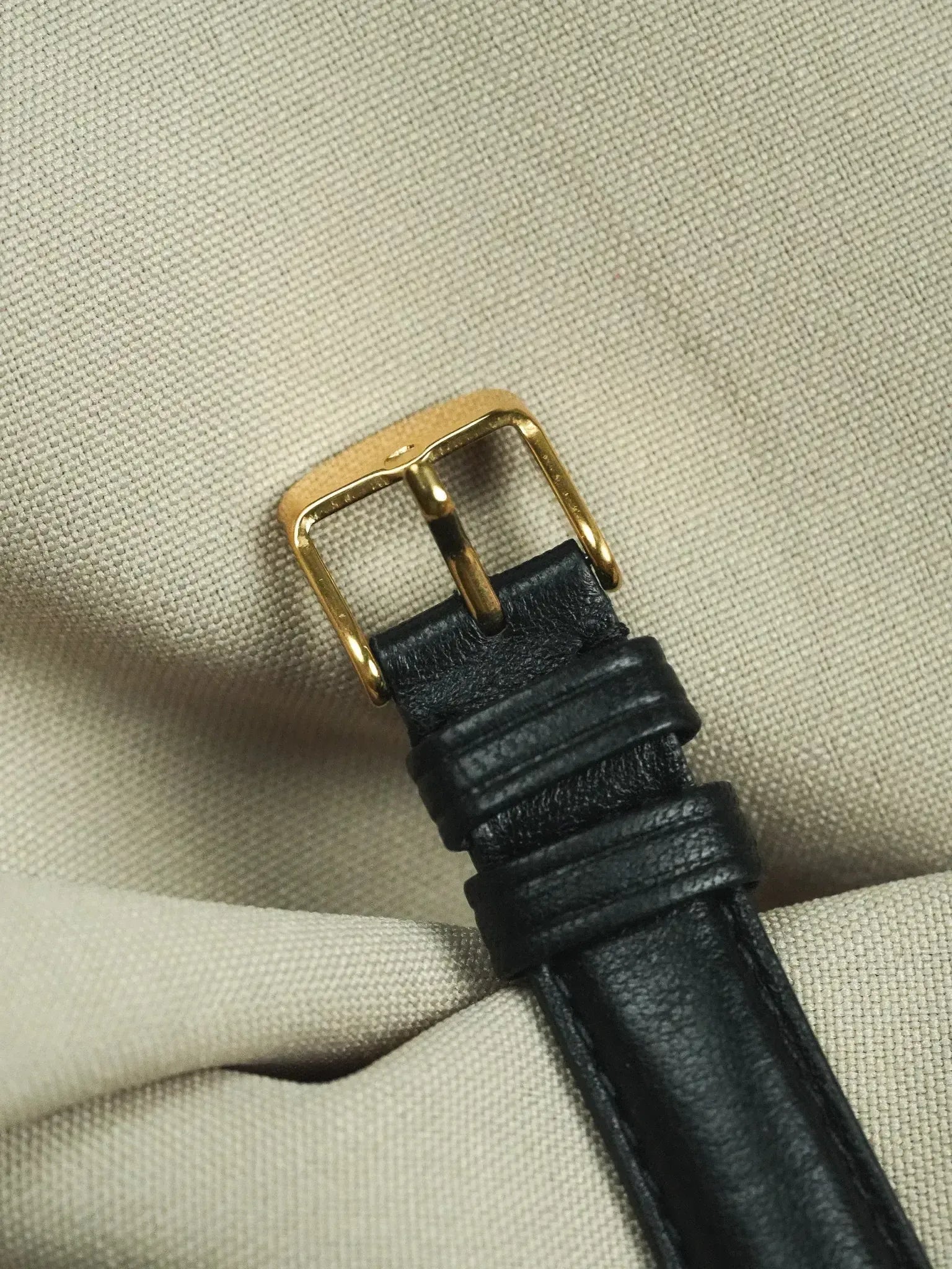 Black leather watch strap with gold buckle on textured fabric