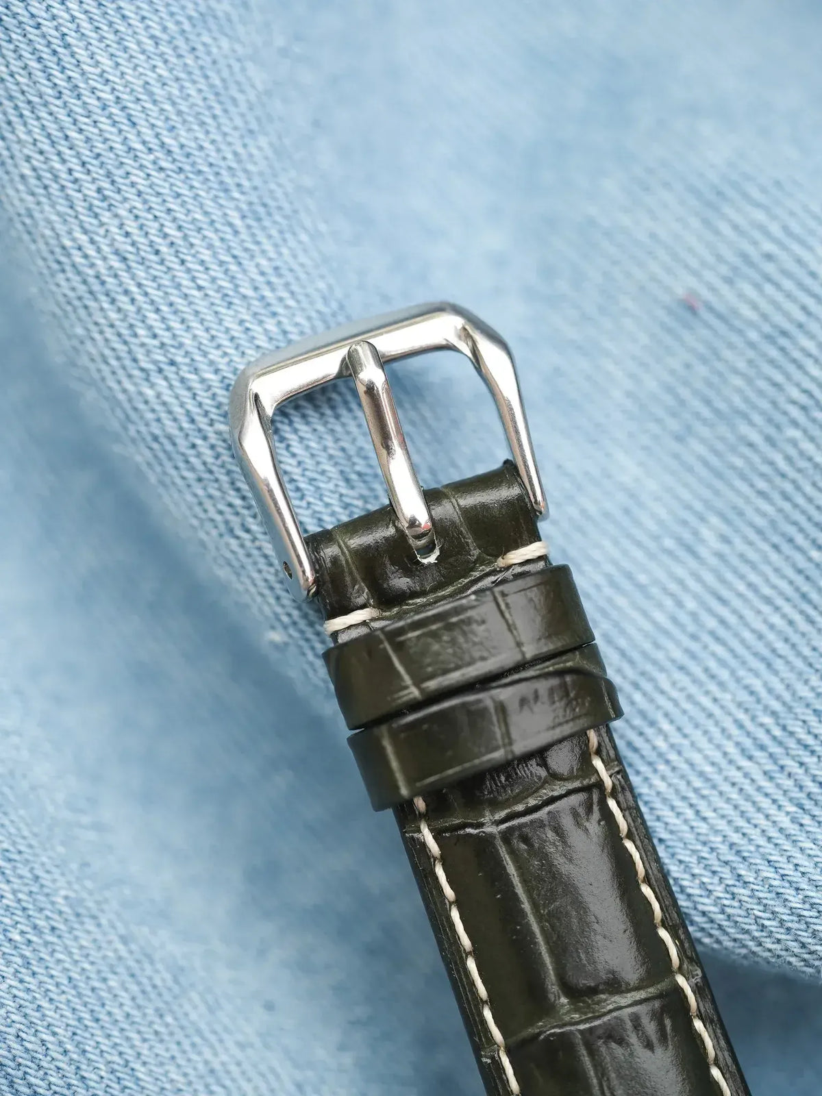 Vintage watch strap with green alligator leather, white stitching, and silver buckle on denim