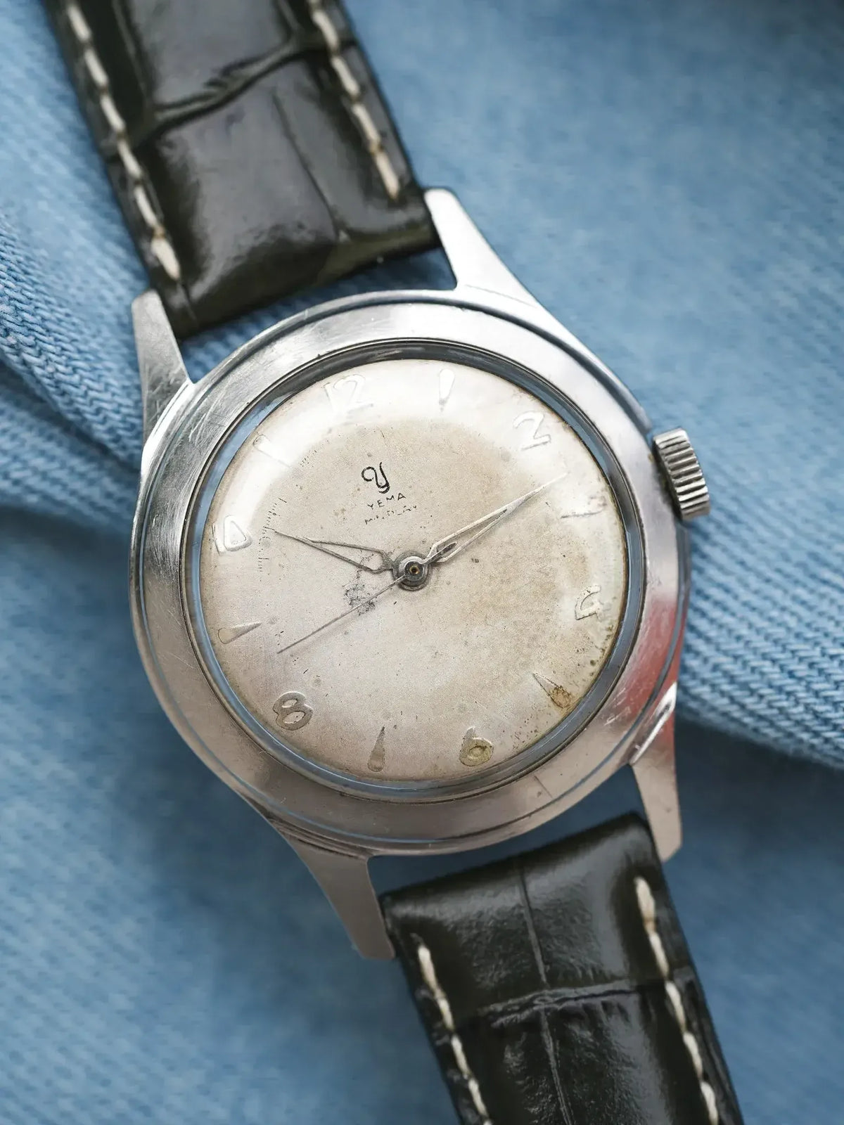 Vintage Yema watch with silver dial, black leather strap, on blue fabric