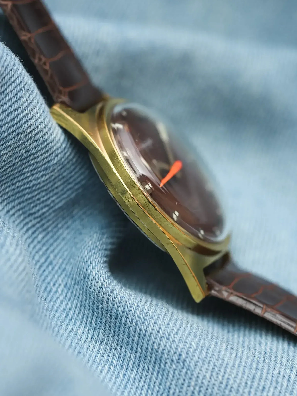 Vintage watch with gold case, brown dial, orange second hand, crocodile strap on denim