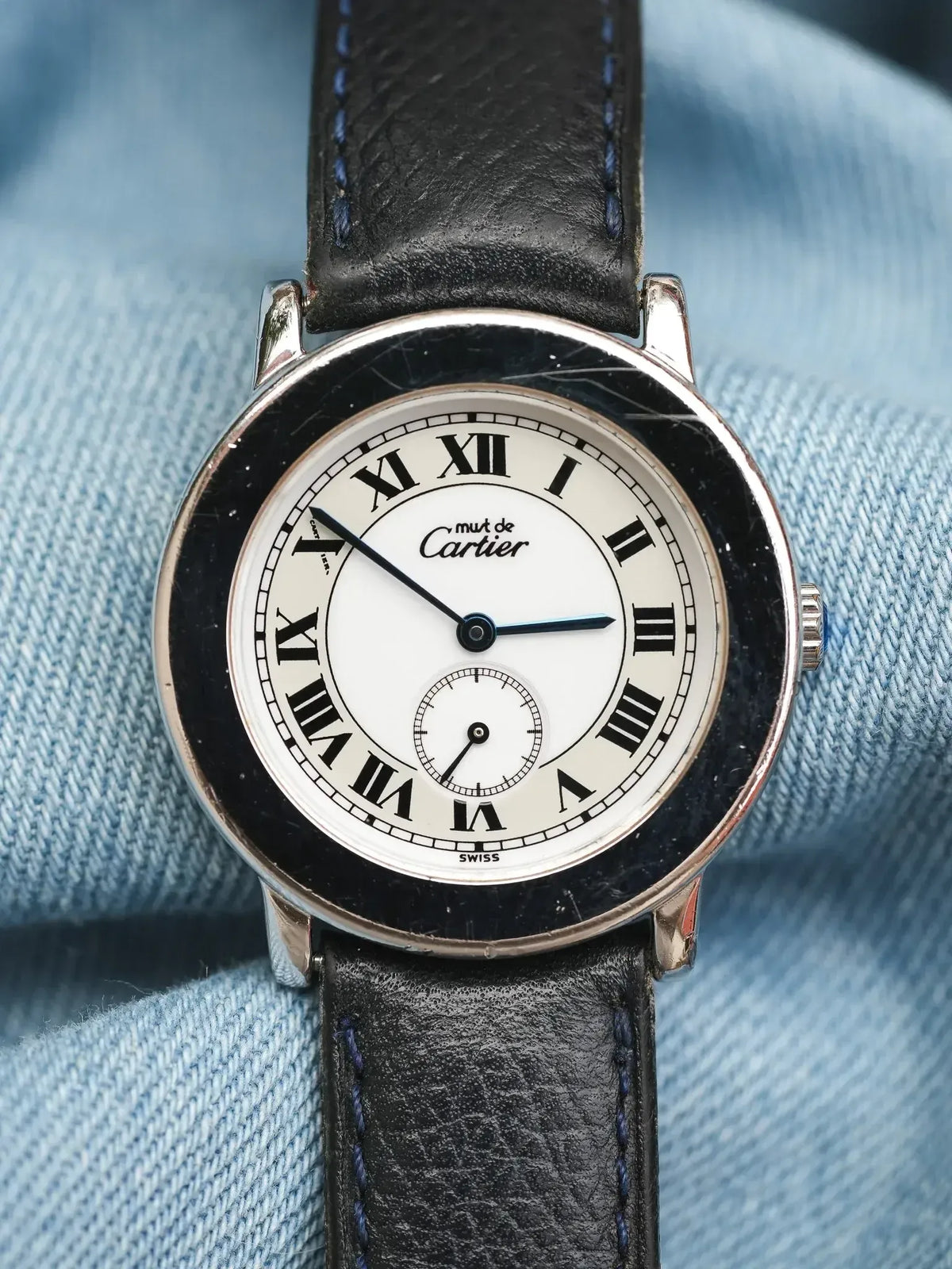 Montre Cartier Must de Cartier vintage, cadran blanc chiffres romains, bracelet cuir noir sur denim