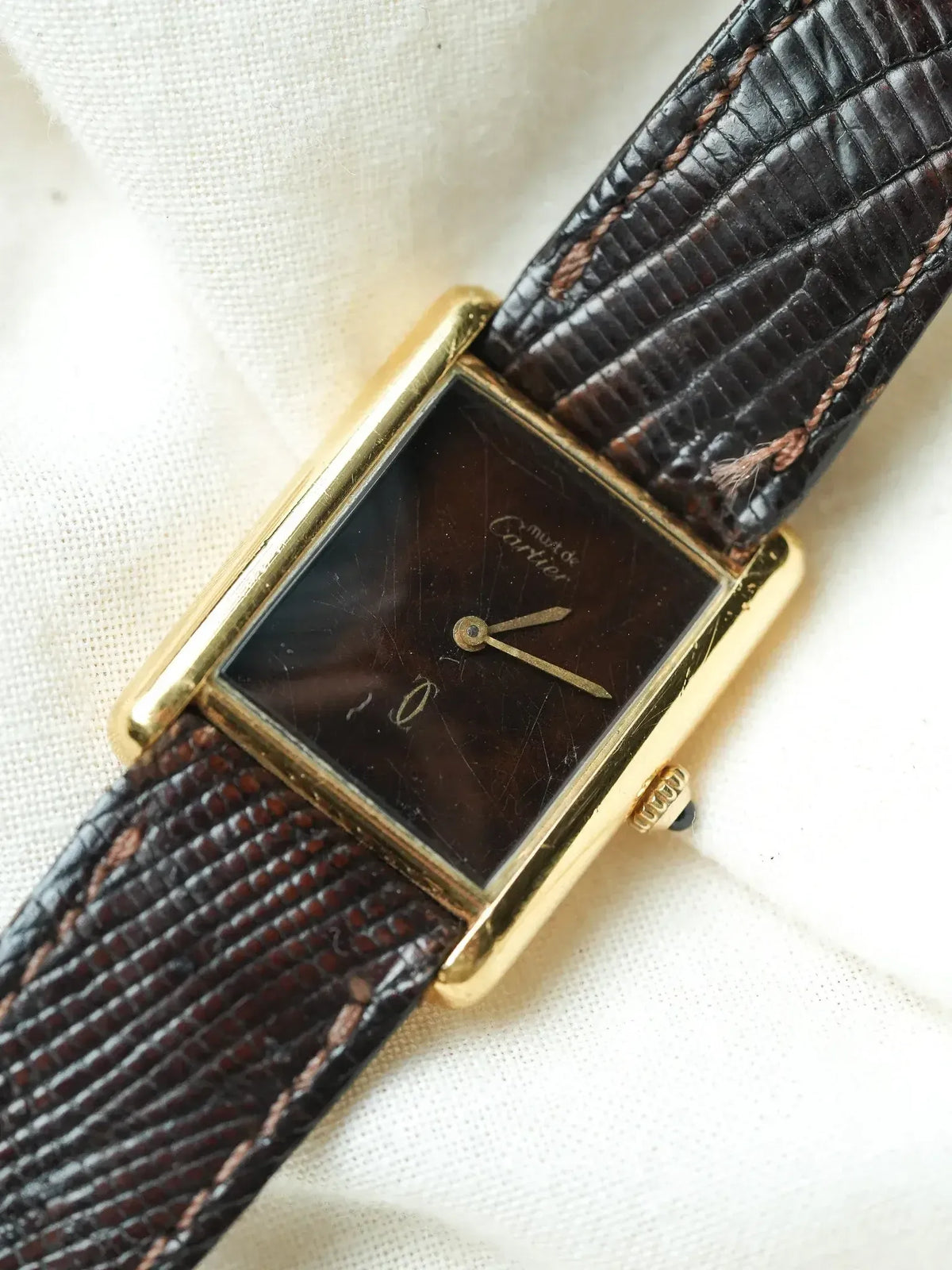 Cartier vintage watch with gold case, dark dial, and brown lizard strap on white fabric