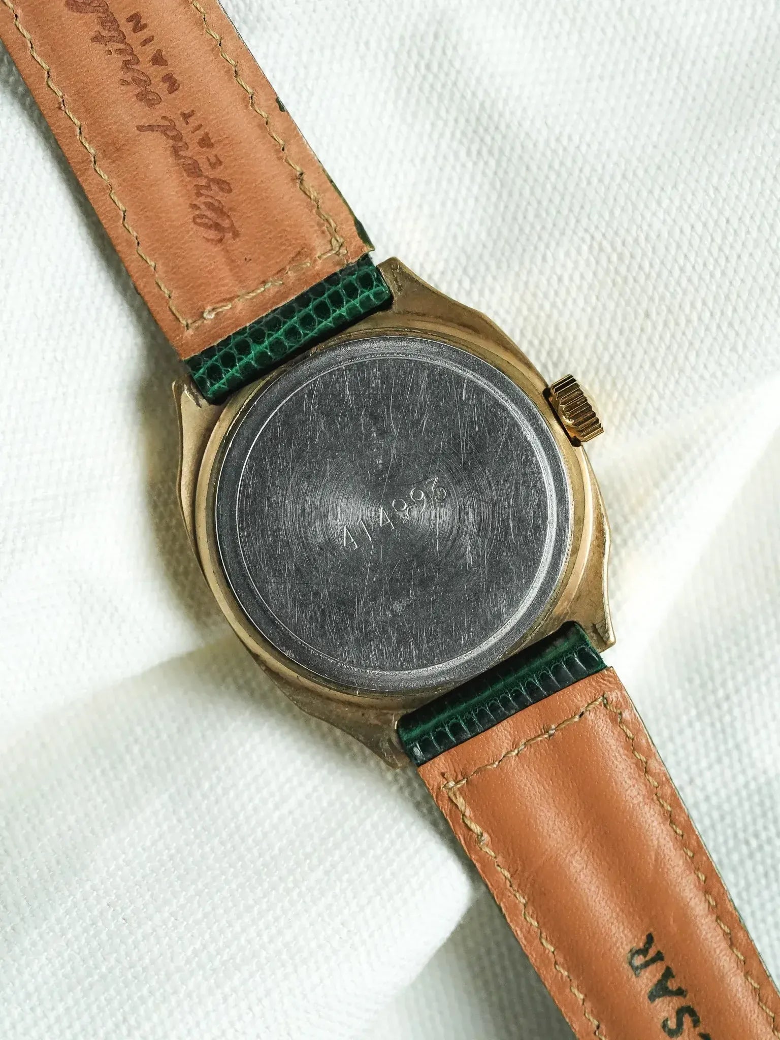Vintage watch with gold case, green strap, and engraved back on white fabric