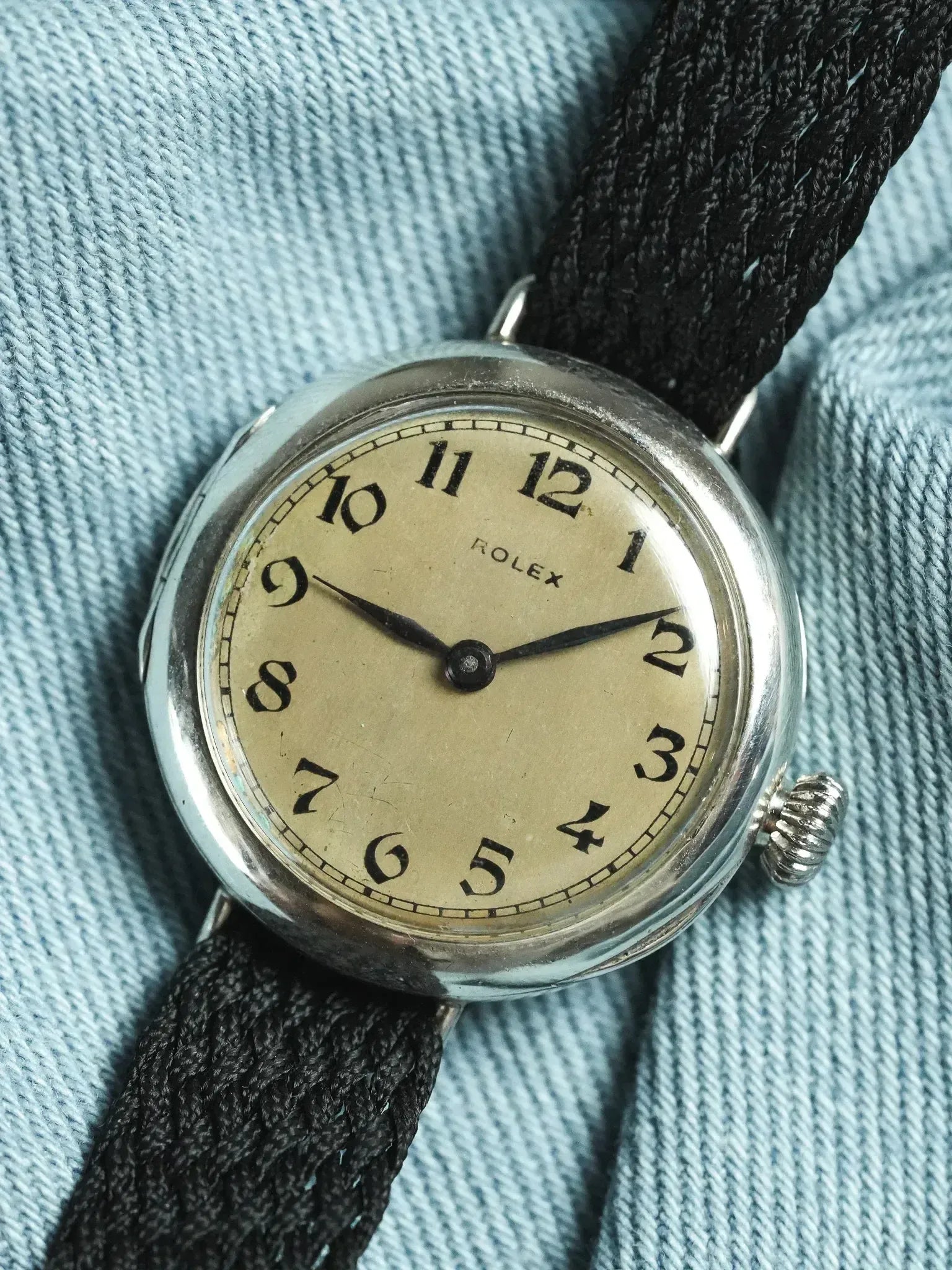 Vintage Rolex wristwatch with cream dial, black numerals, and black fabric strap on denim