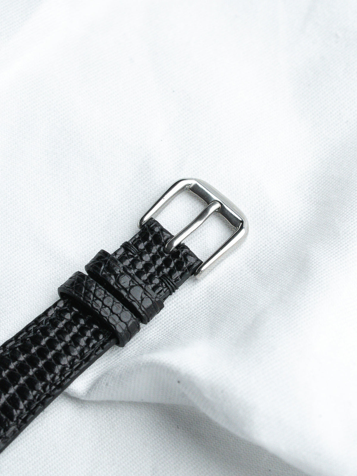 Black lizard skin watch strap with silver buckle on white fabric background