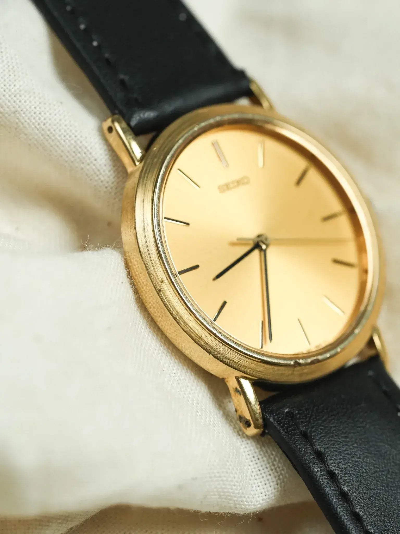 Seiko vintage watch with gold case, cream dial, black leather strap on fabric