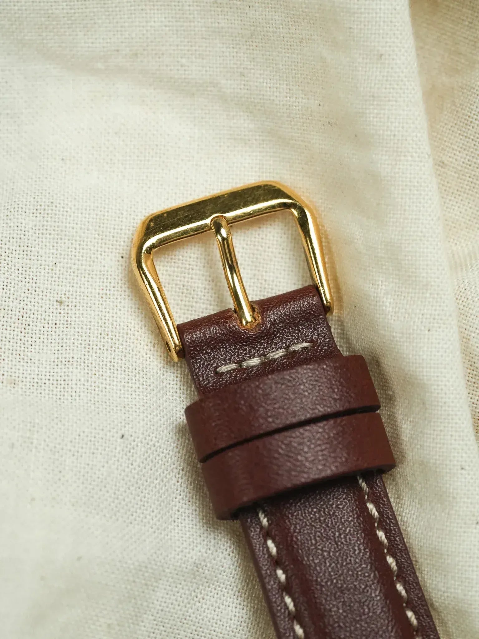 Vintage leather watch strap with gold buckle and white stitching on beige fabric