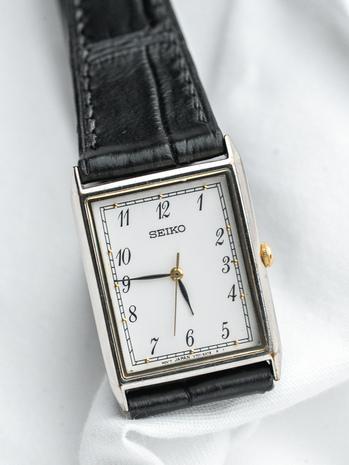 Seiko montre vintage rectangulaire avec cadran blanc, bracelet en cuir noir, accents dorés sur fond blanc