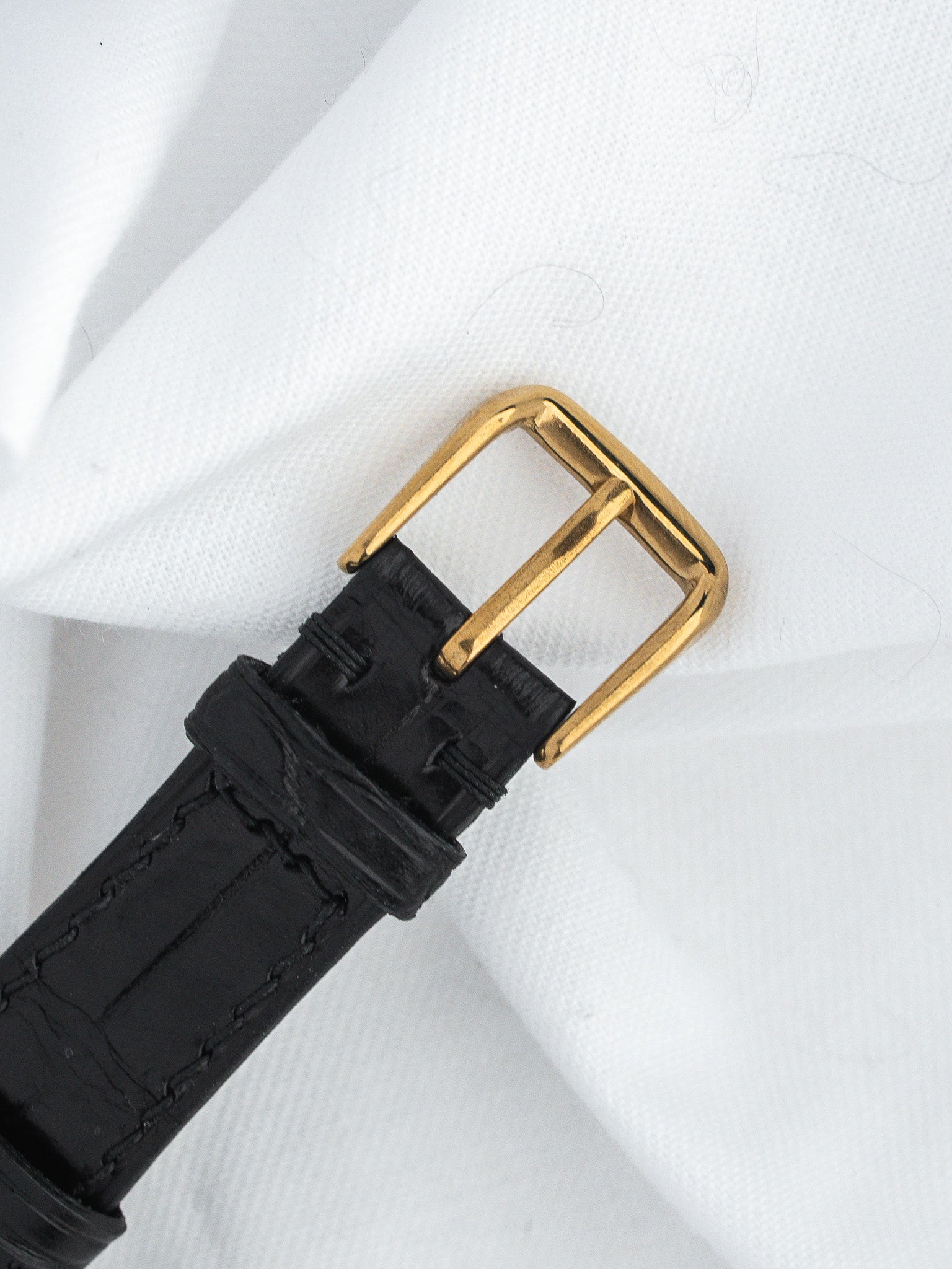 Black leather watch strap with gold buckle, textured stitching, on white fabric background