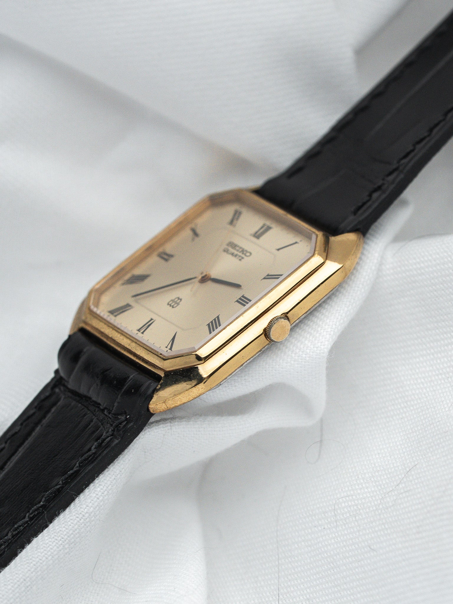 Vintage Seiko quartz watch with gold case, cream dial, Roman numerals, black leather strap on white fabric