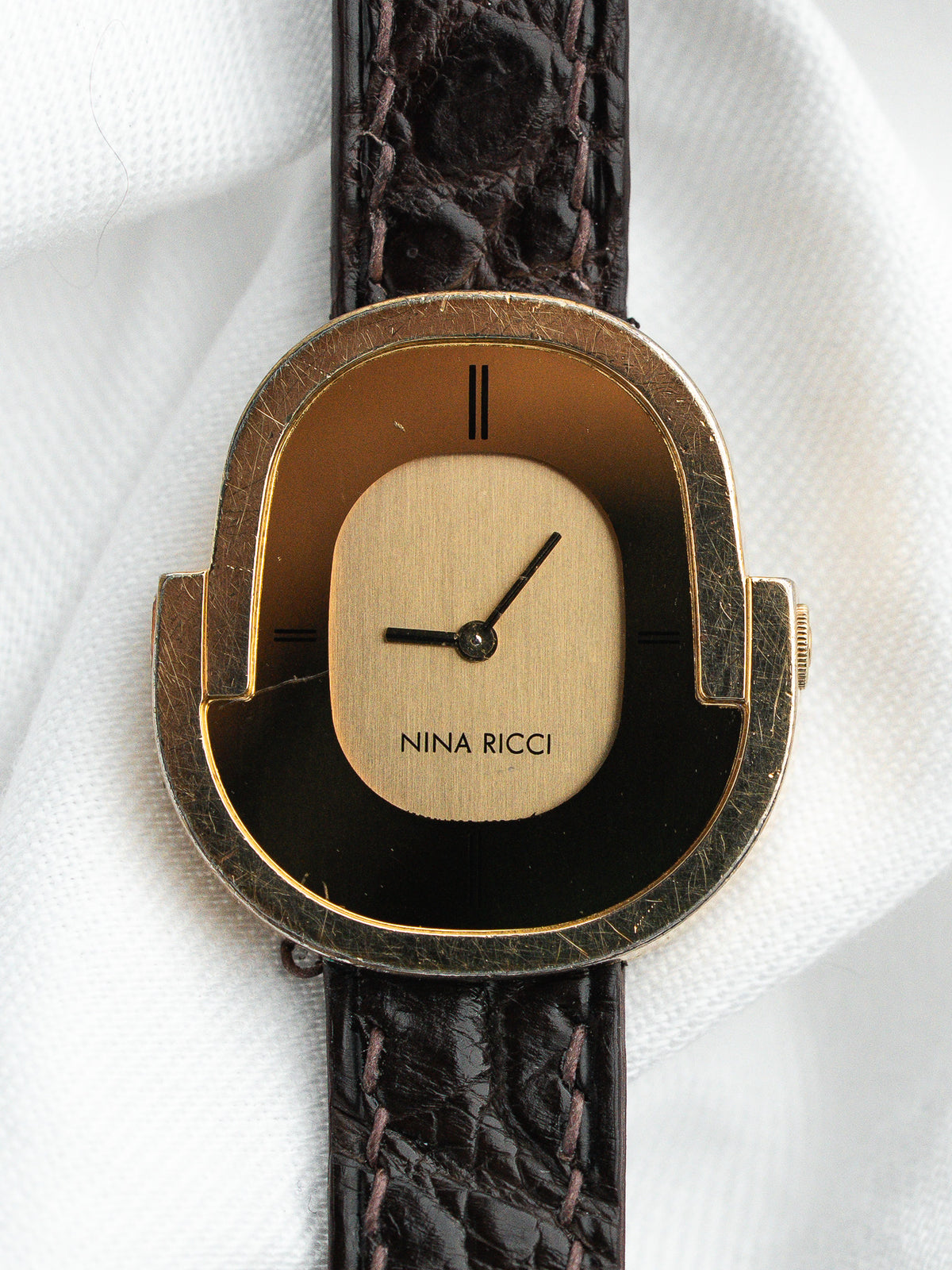 Montre Nina Ricci vintage avec boîtier doré et bracelet en cuir brun texturé.