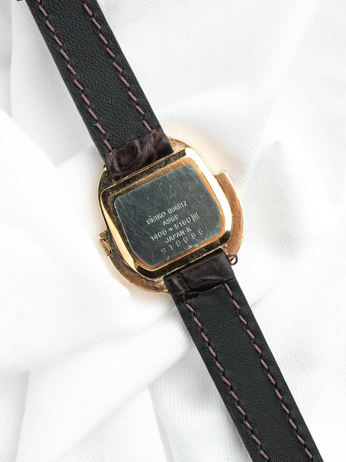 Dos d'une montre Seiko Quartz dorée avec bracelet en cuir noir sur tissu blanc.