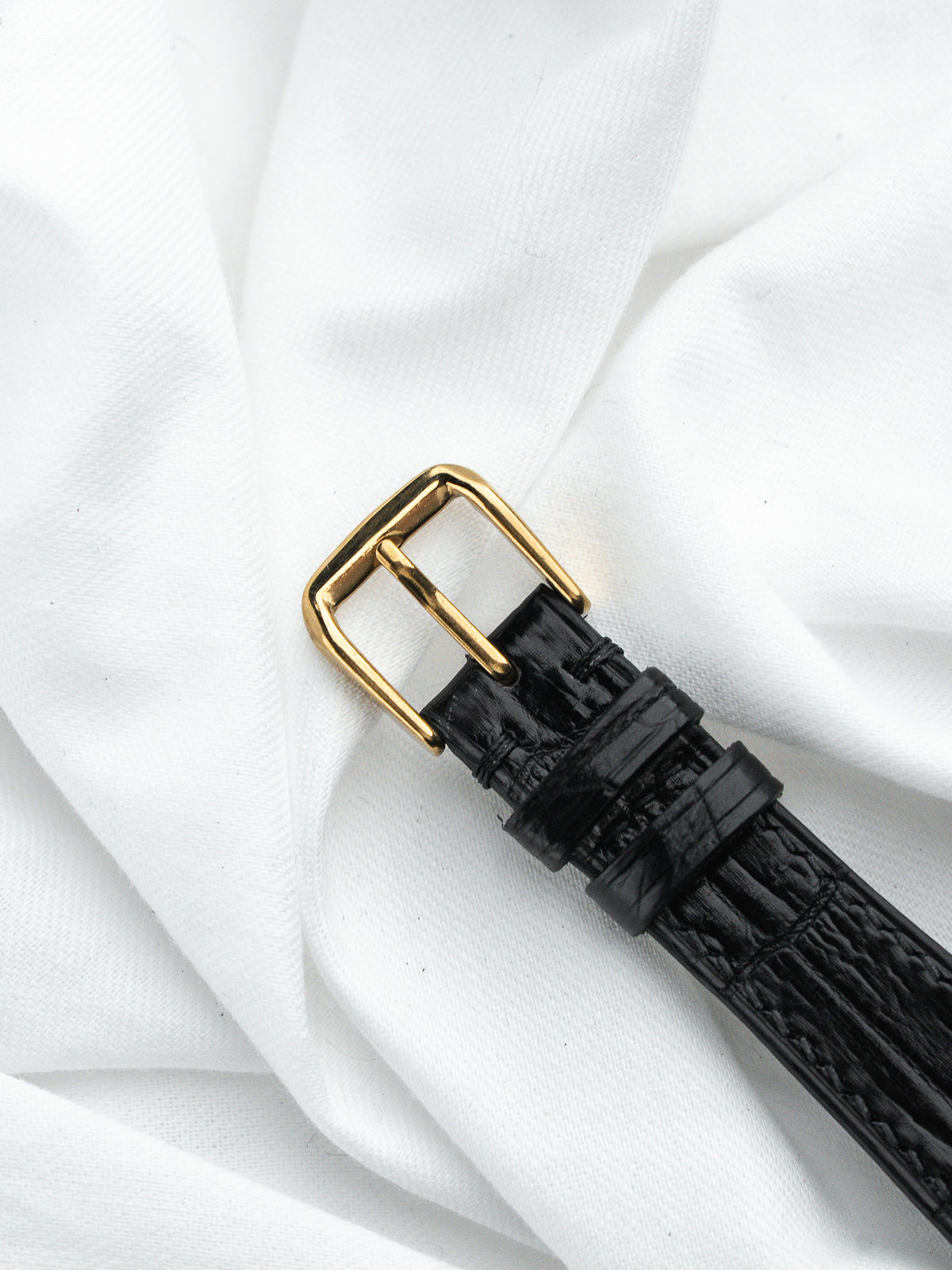 Bracelet de montre en cuir noir avec boucle dorée sur tissu blanc froissé.