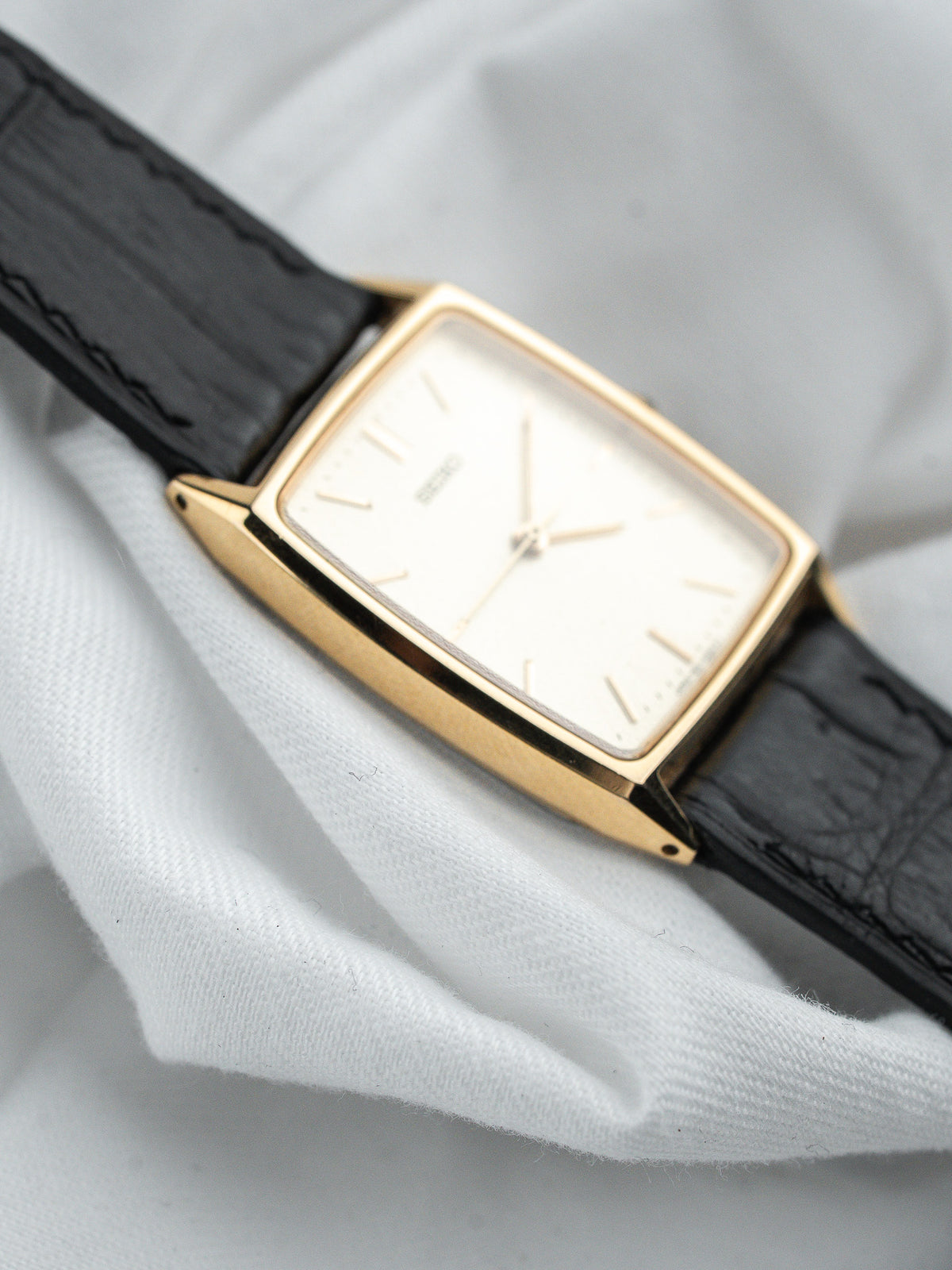 Montre vintage rectangulaire en or avec cadran blanc et bracelet en cuir noir.
