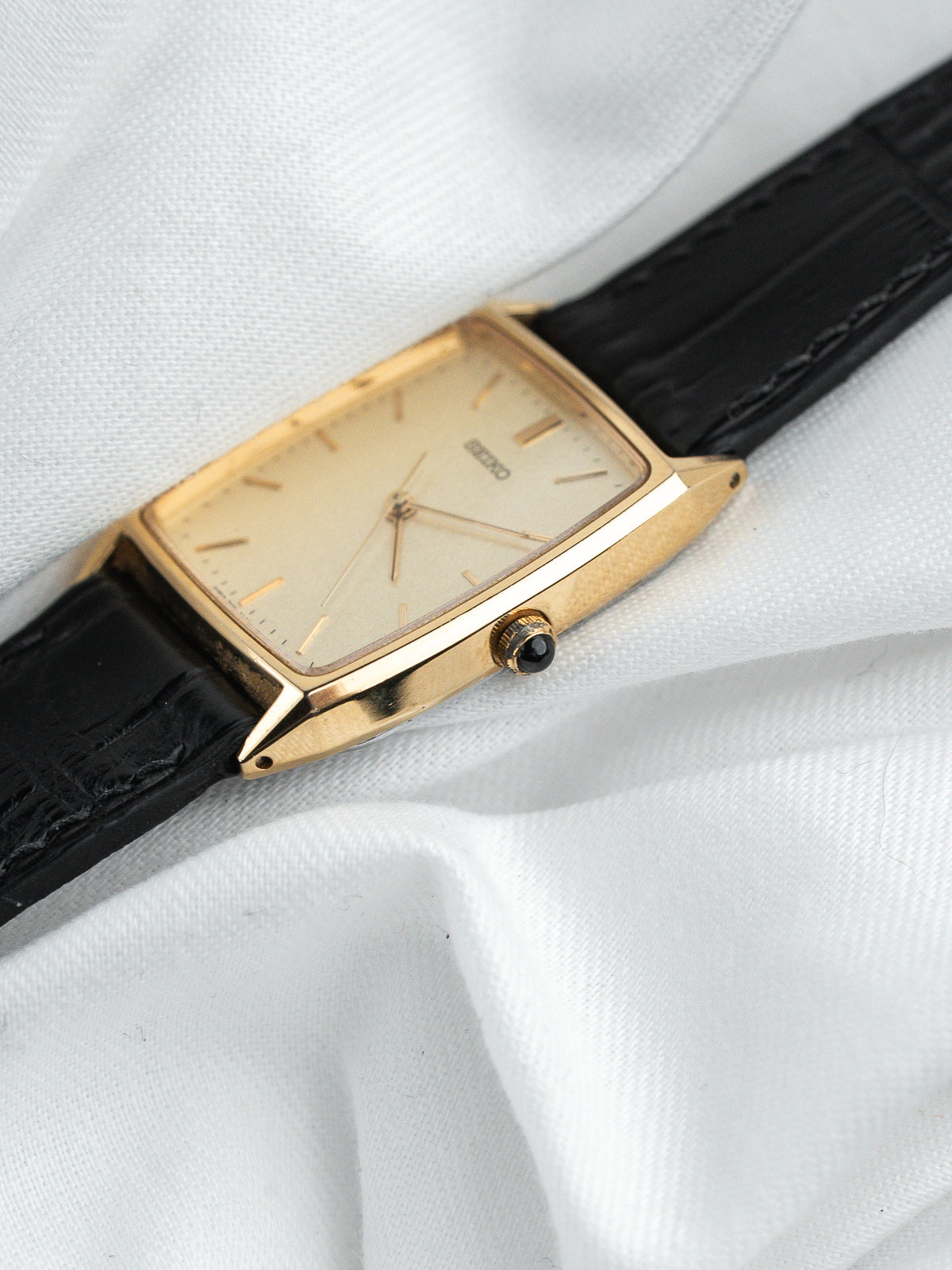 Montre Seiko vintage rectangulaire dorée avec cadran clair et bracelet en cuir noir.