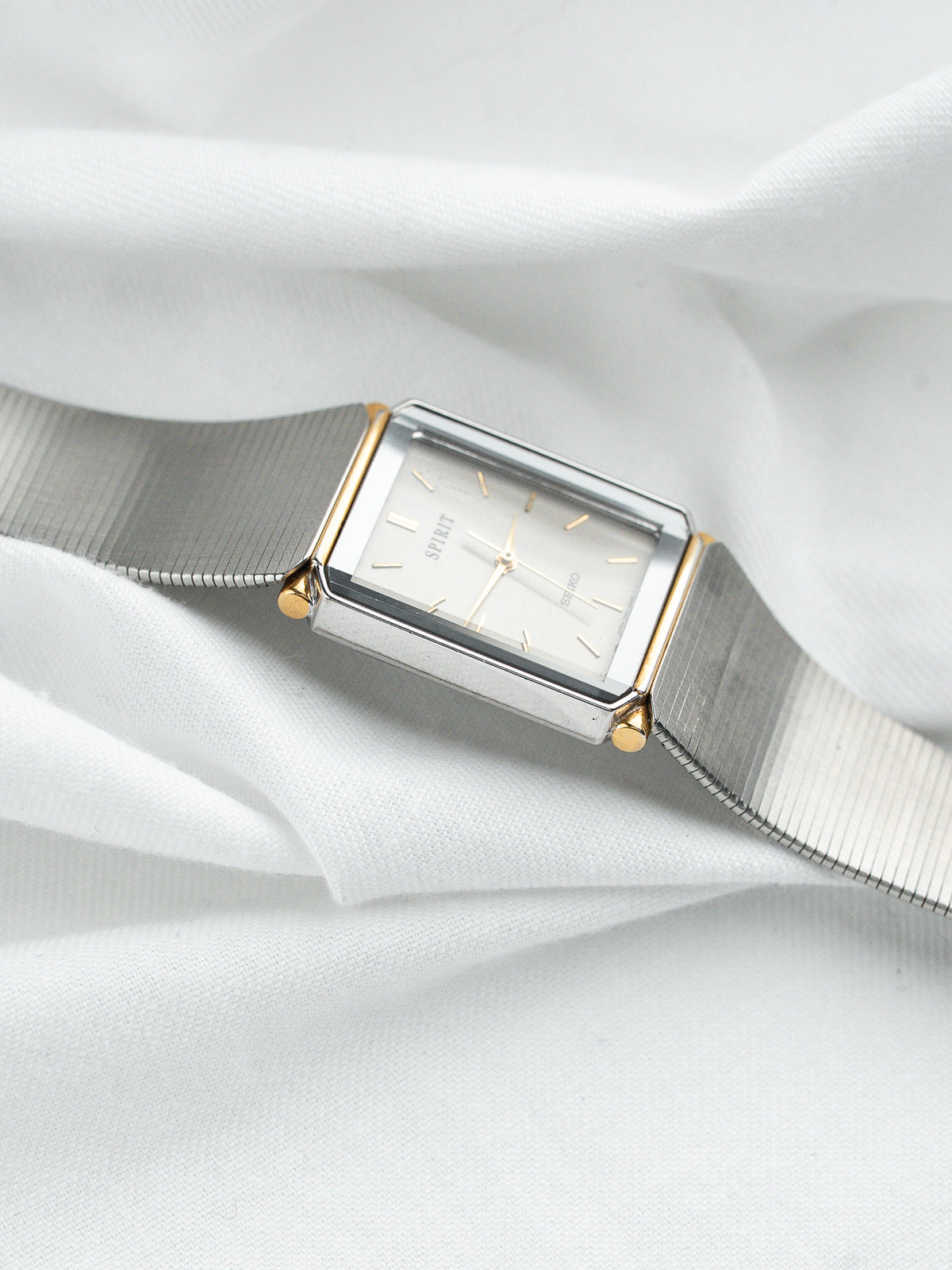 Montre vintage rectangulaire en acier argenté avec détails dorés sur fond de tissu blanc.